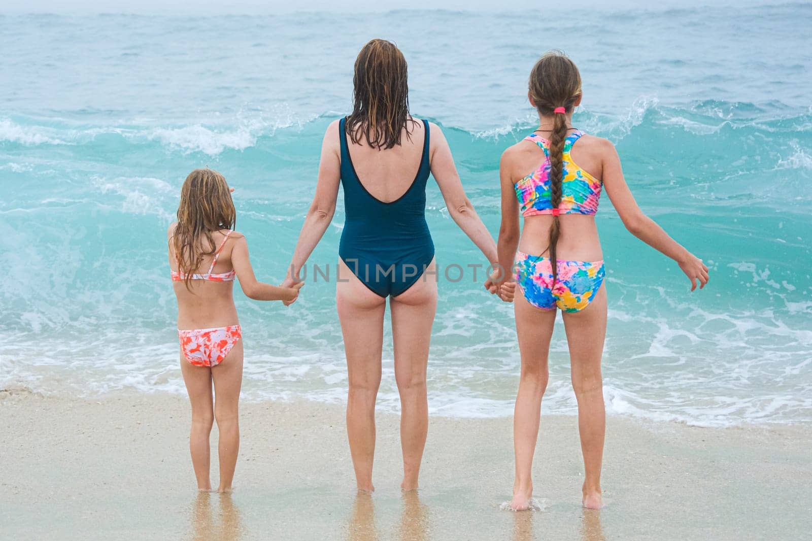 Mother and children playing on the ocean beach. Family enjoying the ocean. Mother holds girls's hands and they all look at the ocean together by esvetleishaya