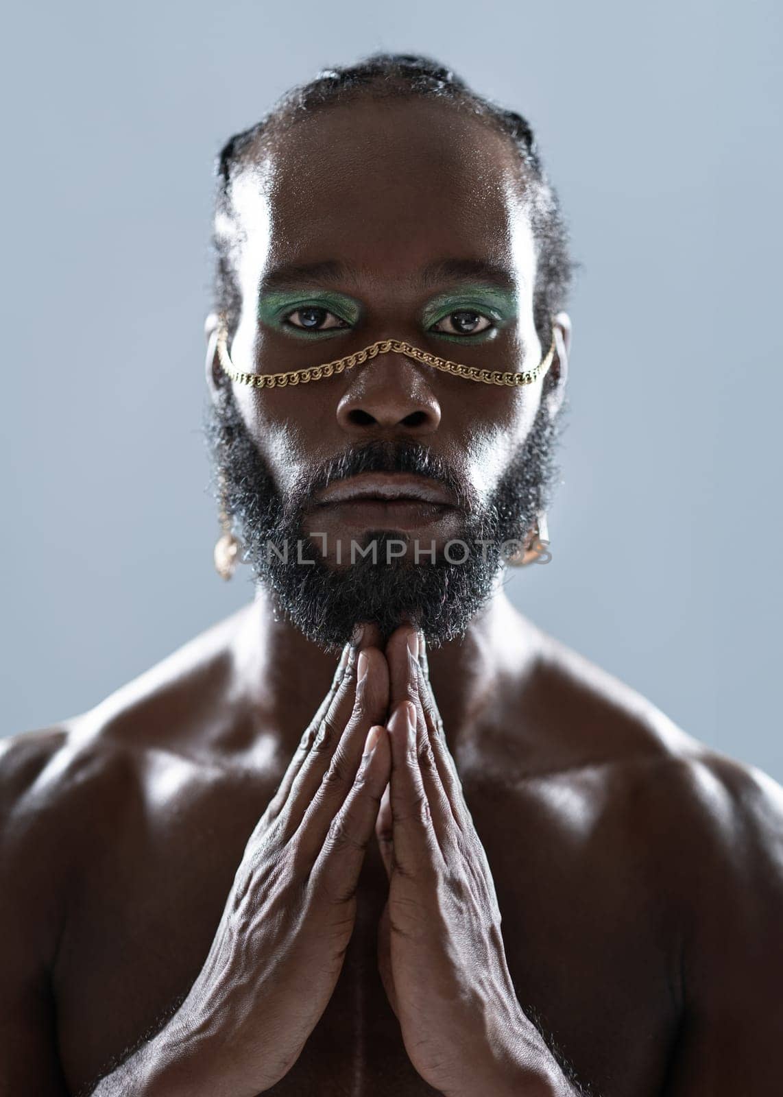 Portrait of serious African American gay man with blue eyeshadows and golden accessory on face. Shirtless adult transgender joining hands and looking at camera on blue background.