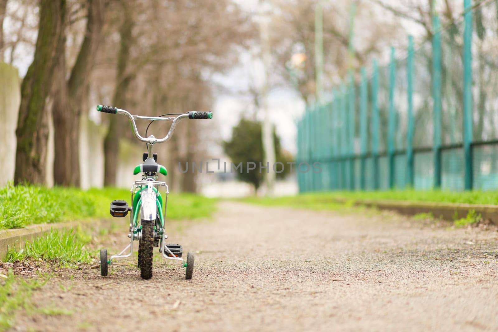 A green bicycle for children with additional wheels at road in park. Copy space