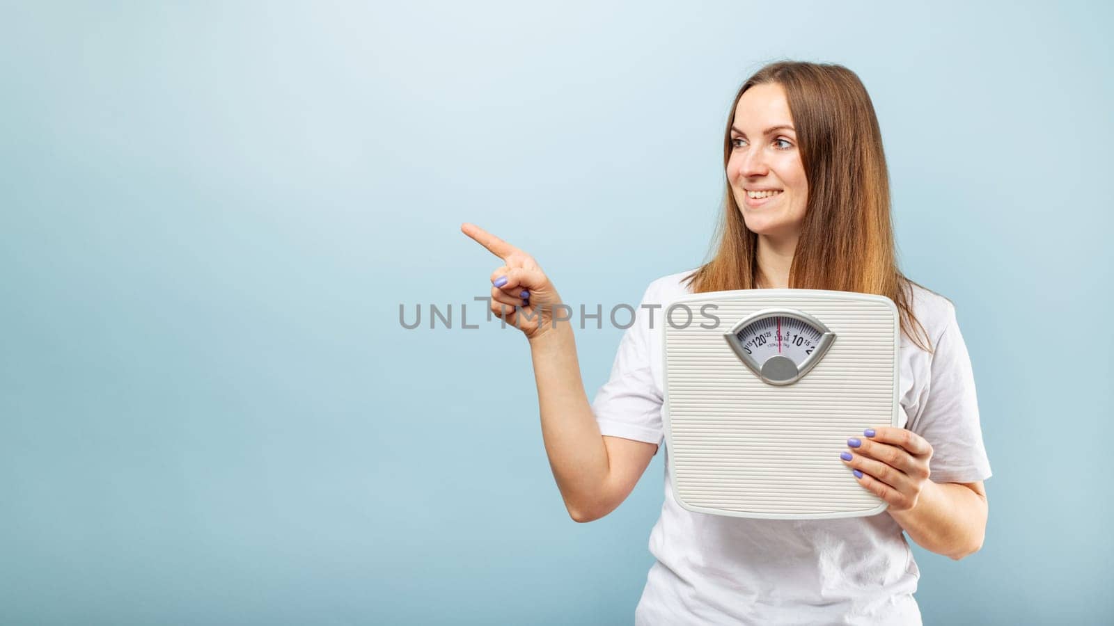 Young brunette woman with weighing scale pointing with finger to the side on blue background