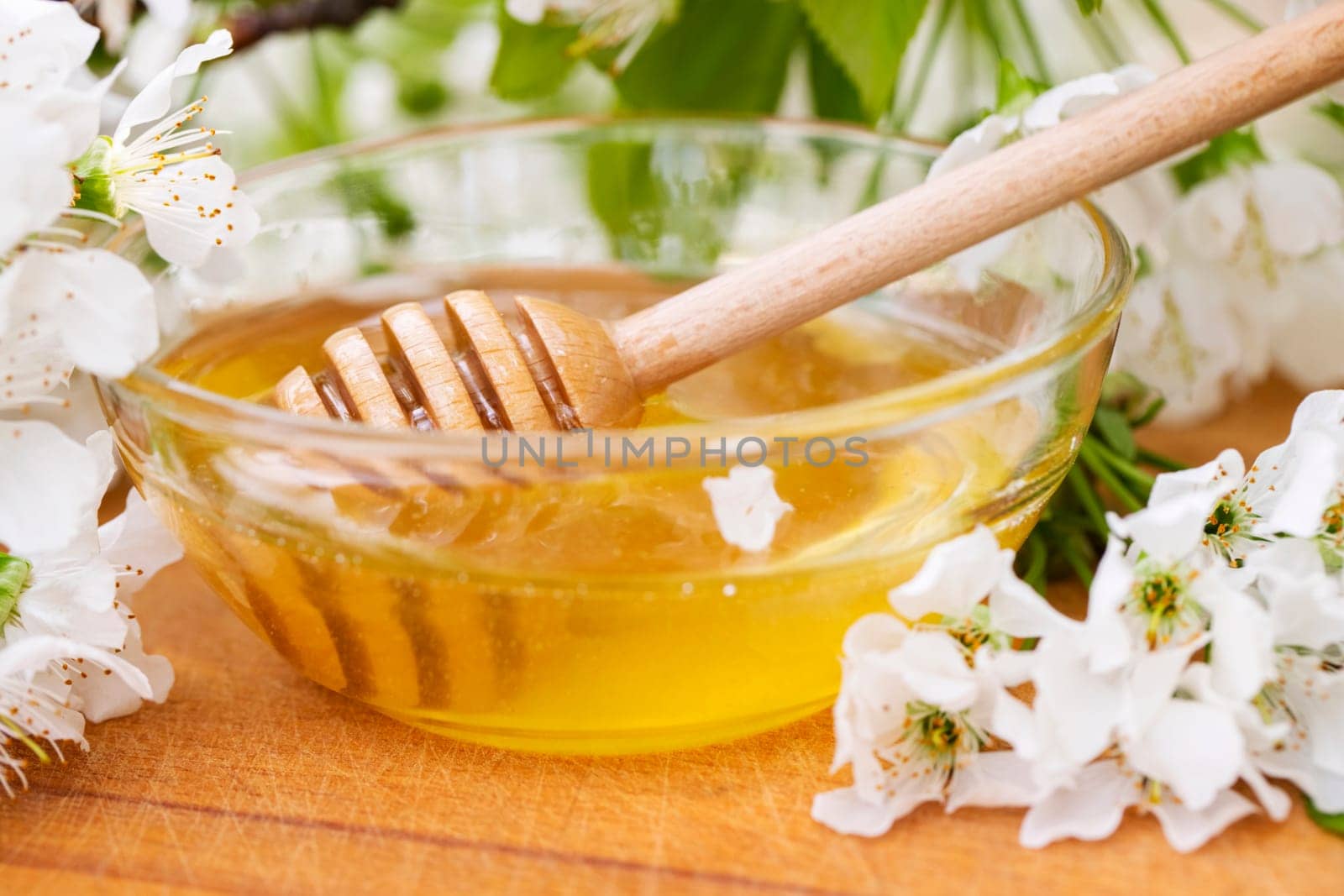 Flower honey in a glass bowl with wooden honey spoon on wooden table with blossoming flowers.