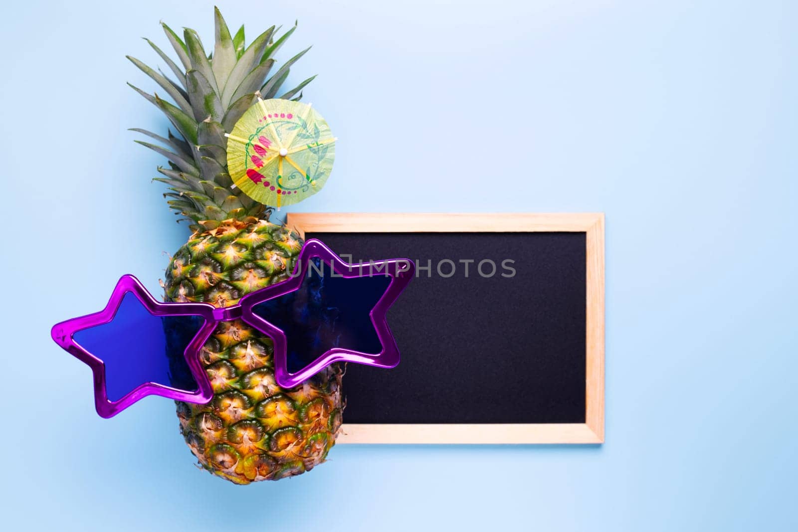 Pineapple in sunglasses with empty copy space blackboard for your text on blue background. Summer vacation party time concept.