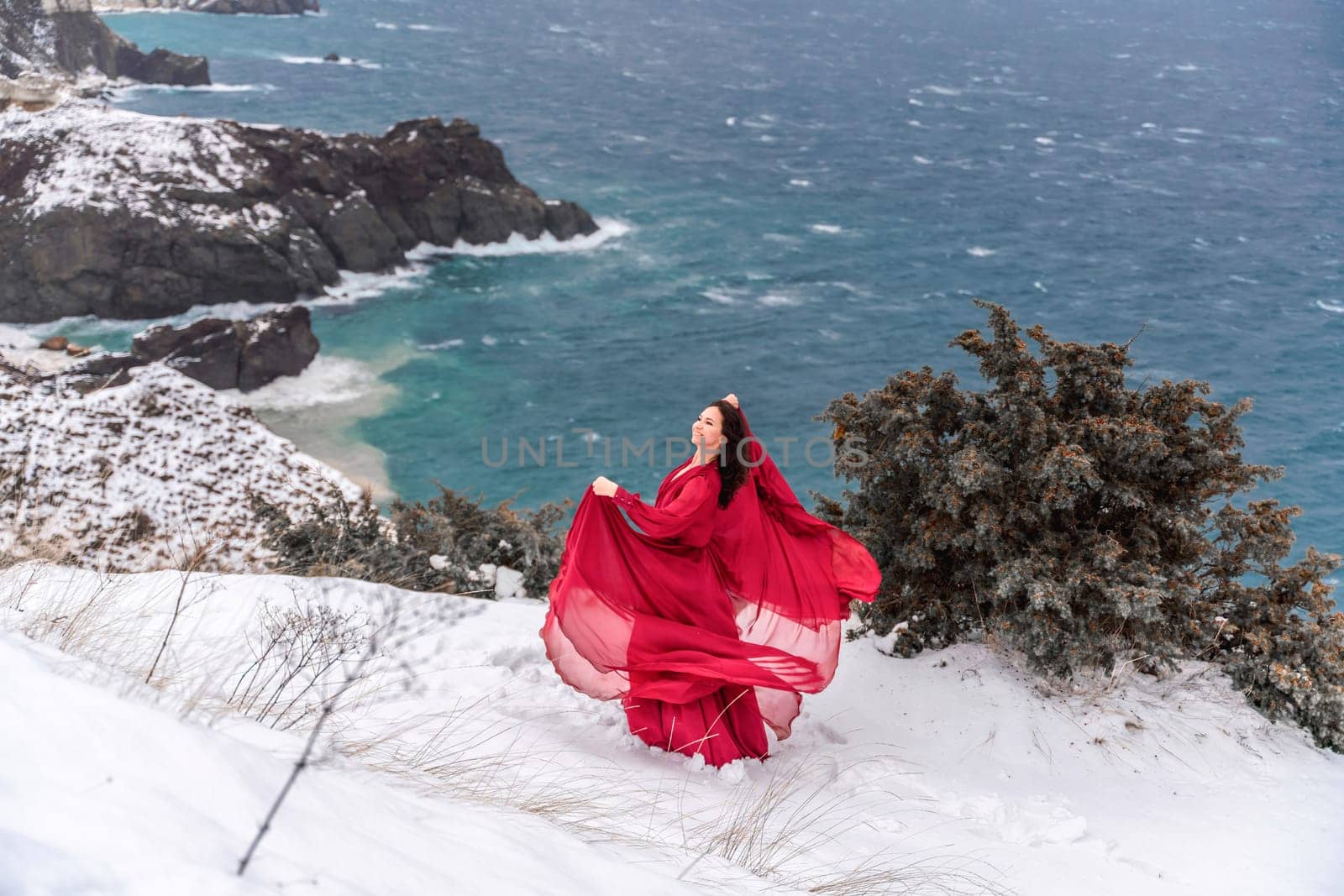 Woman red dress snow sea. Happy woman in a red dress in the snowy mountains by the emerald sea. The wind blows her clothes, posing against sea and snow background