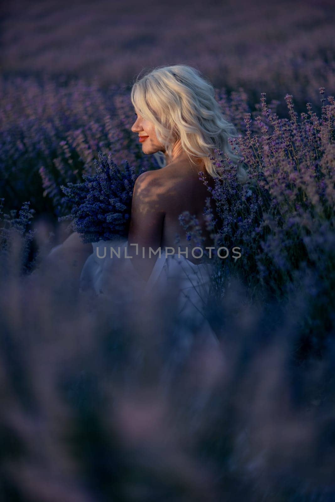 Blonde woman poses in lavender field at sunset. Happy woman in white dress holds lavender bouquet. Aromatherapy concept, lavender oil, photo session in lavender.