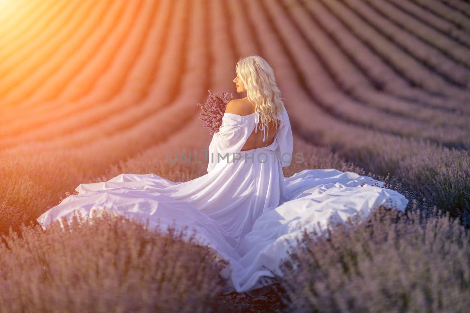 Blonde woman poses in lavender field at sunset. Happy woman in white dress holds lavender bouquet. Aromatherapy concept, lavender oil, photo session in lavender.