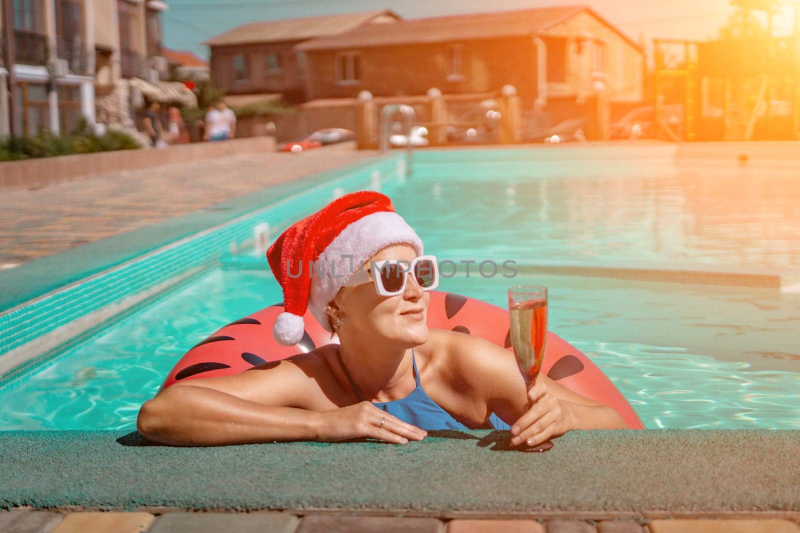 Woman pool Santa hat. A happy woman in a blue bikini, a red and white Santa hat and sunglasses poses near the pool with a glass of champagne standing nearby. Christmas holidays concept. by Matiunina