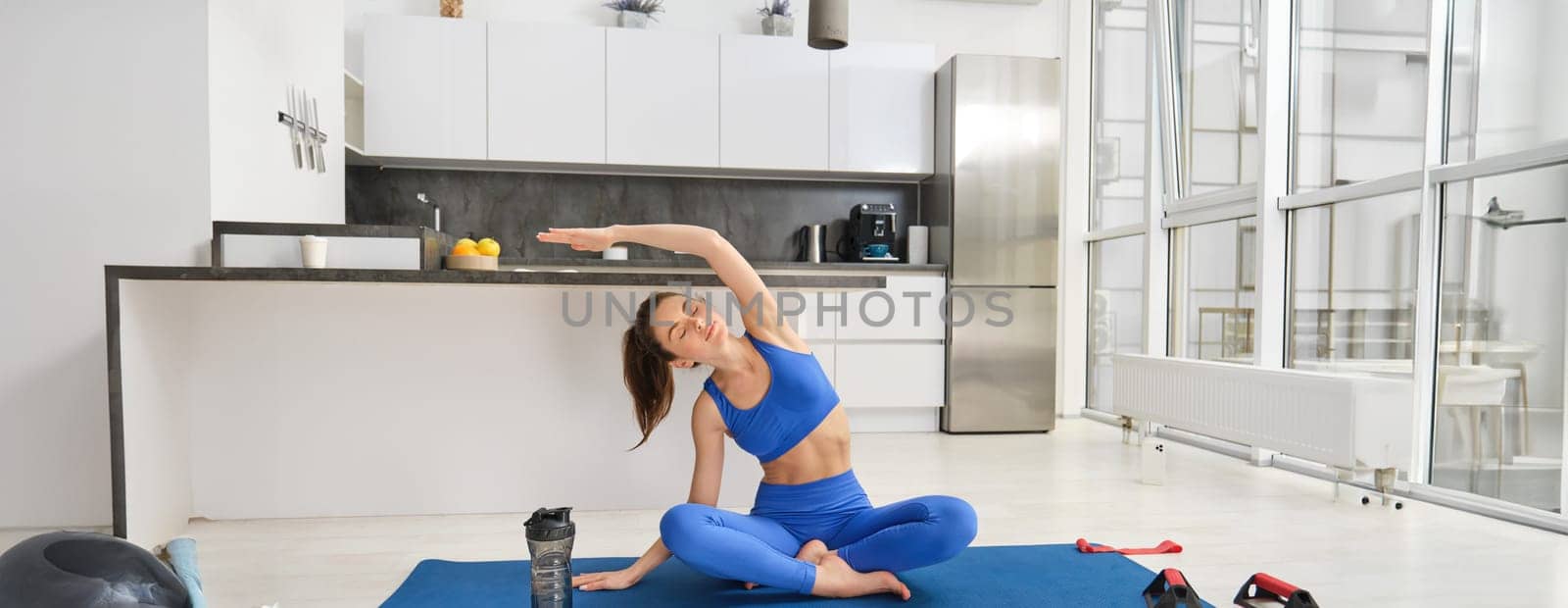 Young sporty woman in fitness clothes, sits on rubber yoga mat, stretches arms sideways, meditates in living room, exercises in blue sportswear by Benzoix