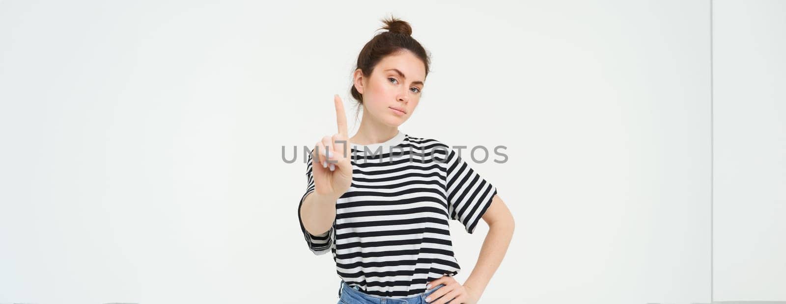Image of confident woman, shows one finger, taboo, stop gesture, disapprove smth, gives warning, stands over white background.