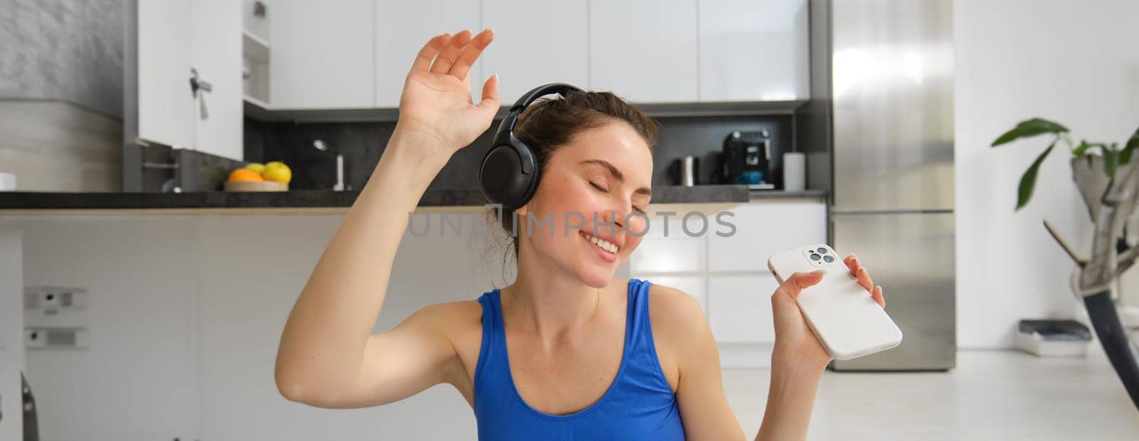 Portrait of fitness instructor, woman with headphones and smartphone, dancing at home, workout inside her house, wearing blue sportsbra.