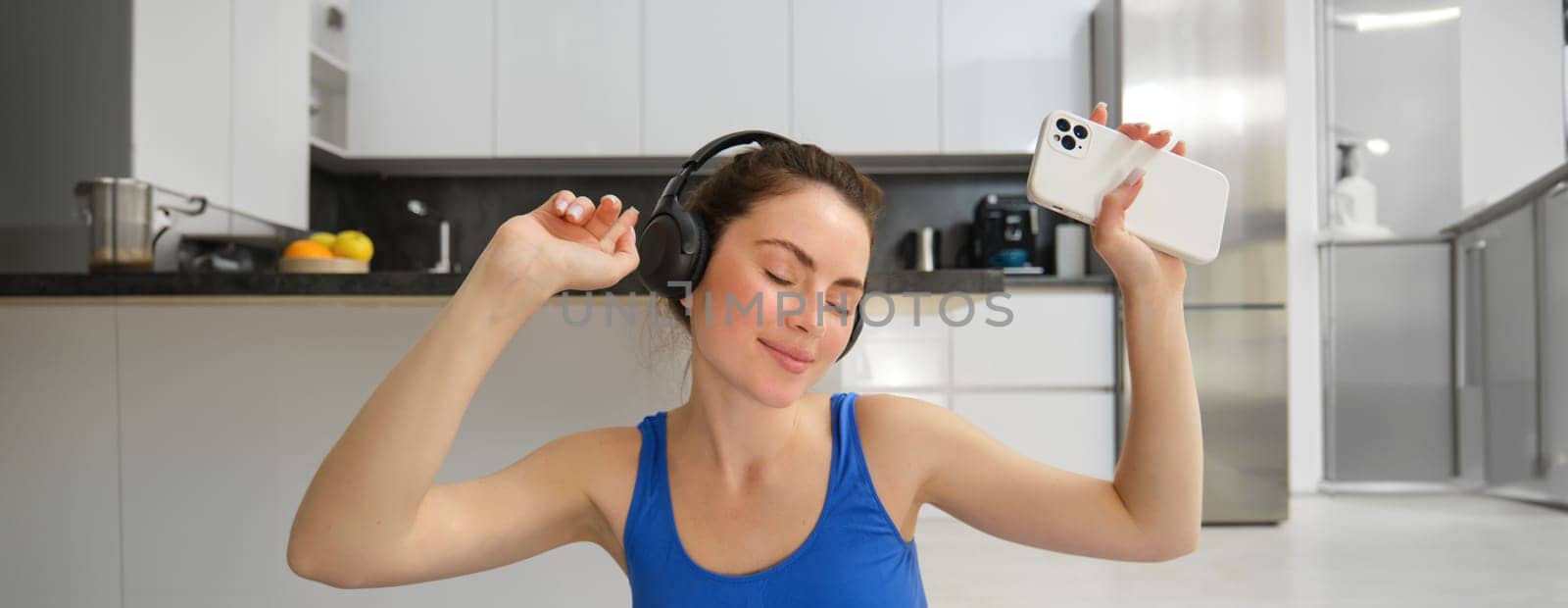 Happiness, sports and wellbeing. Young woman dancing in headphones, holding smartphone, doing workout at home, fitness training inside her house by Benzoix
