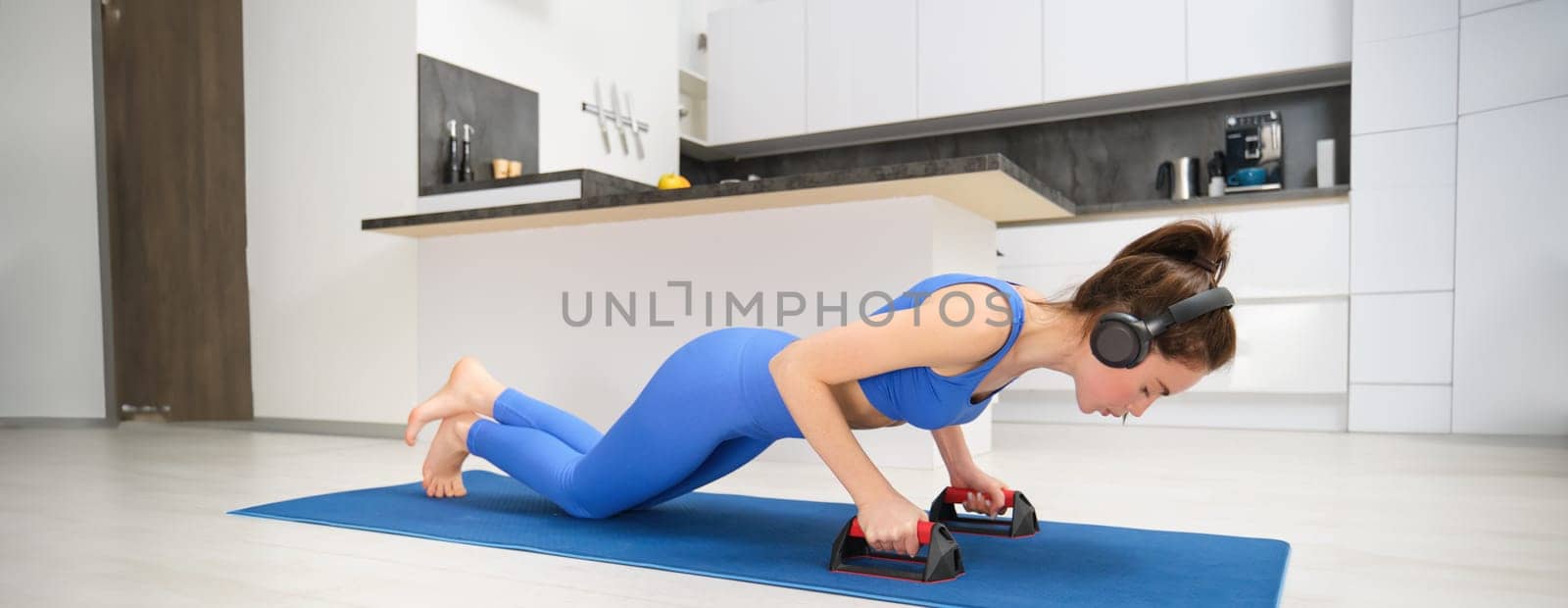 Portrait of fitness woman in leggings, doing push-up with sports equpment, workout from home, standing on yoga mat in living room, listening music in headphones.