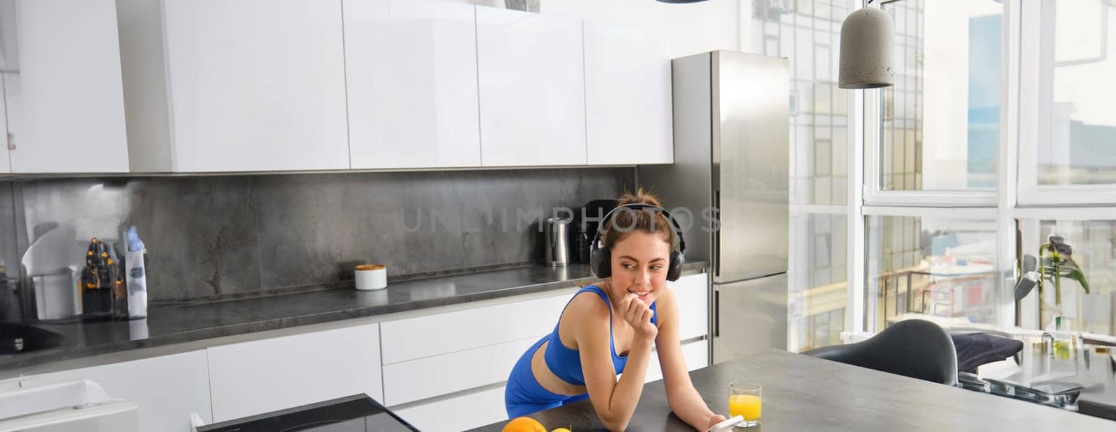 Lifestyle and workout. Young smiling woman in headphones, standing in kitchen with smartphone, drinking orange juice and listening music, heading over to gym, going jogging by Benzoix
