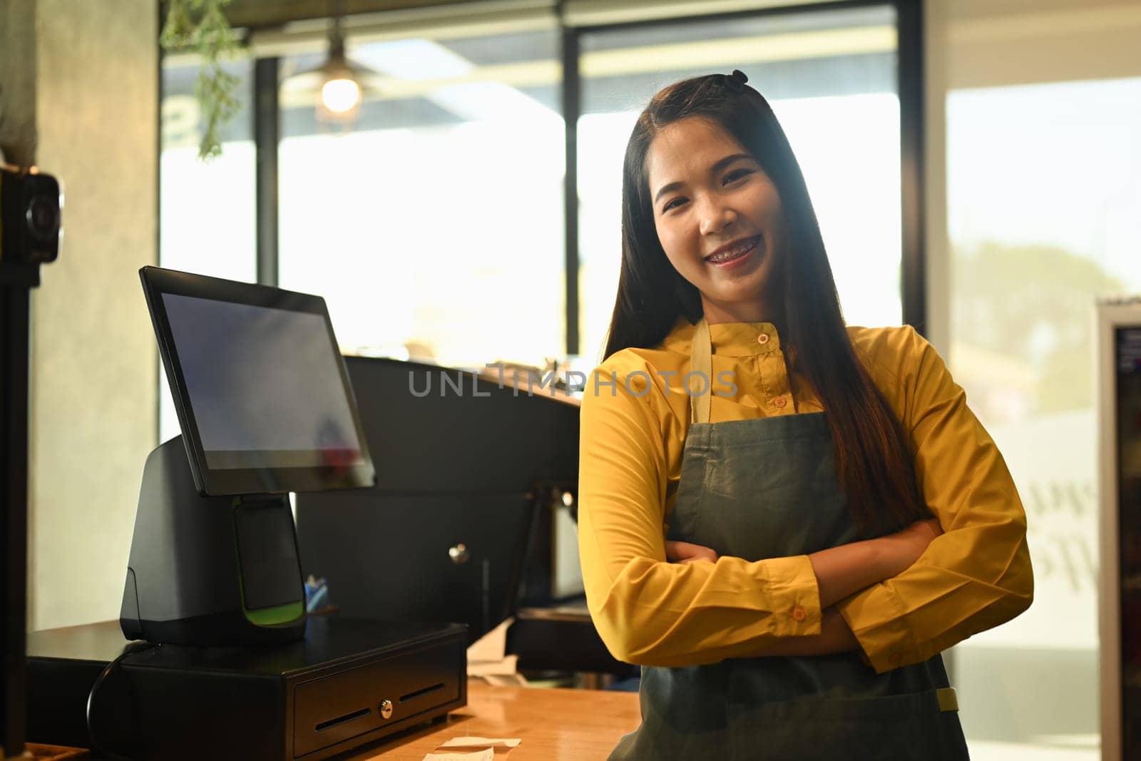 Friendly small coffee shop owner standing with arms crossed behind counter with cash register by prathanchorruangsak