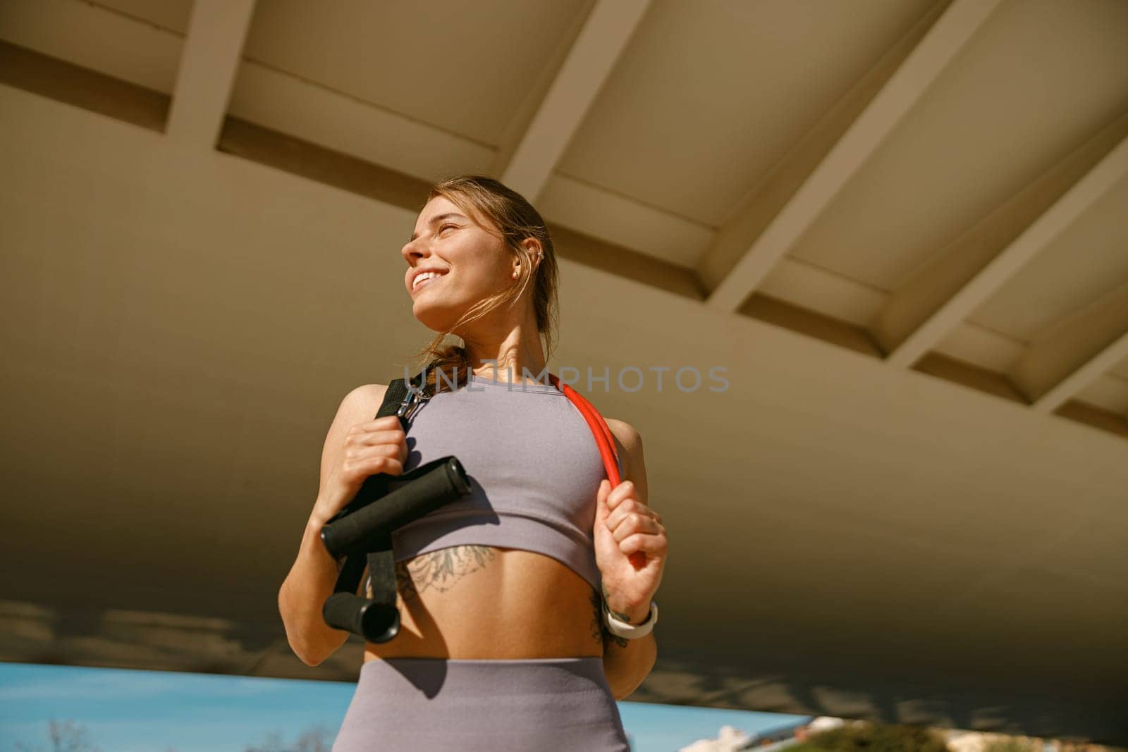Smiling athletic woman in sportswear with a resistance band slung around her neck standing outdoors by Yaroslav_astakhov