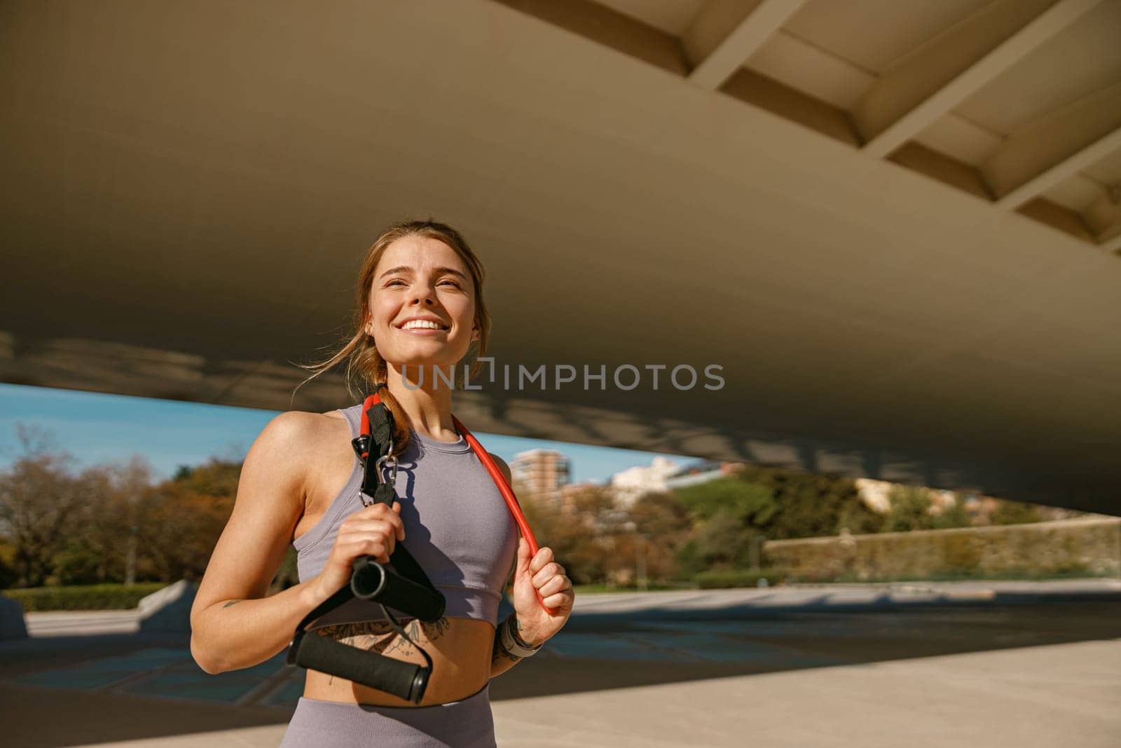 Athletic woman with a resistance band slung around her neck standing outdoors and looks away by Yaroslav_astakhov