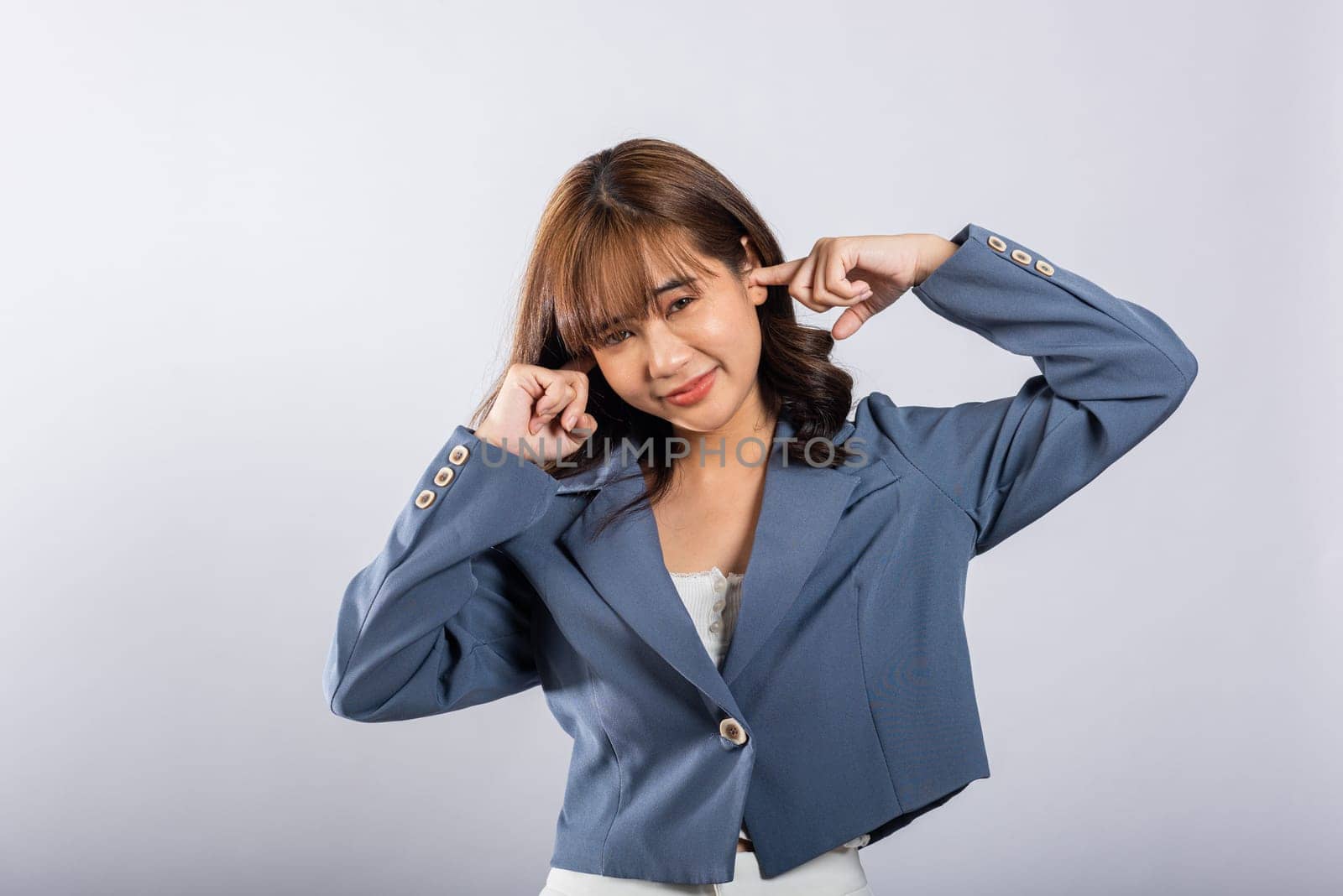 An Asian woman is captured in a studio shot, conveying her stress and discomfort caused by loud noise. She covers her ears and closes her eyes tightly, isolated on a white background.