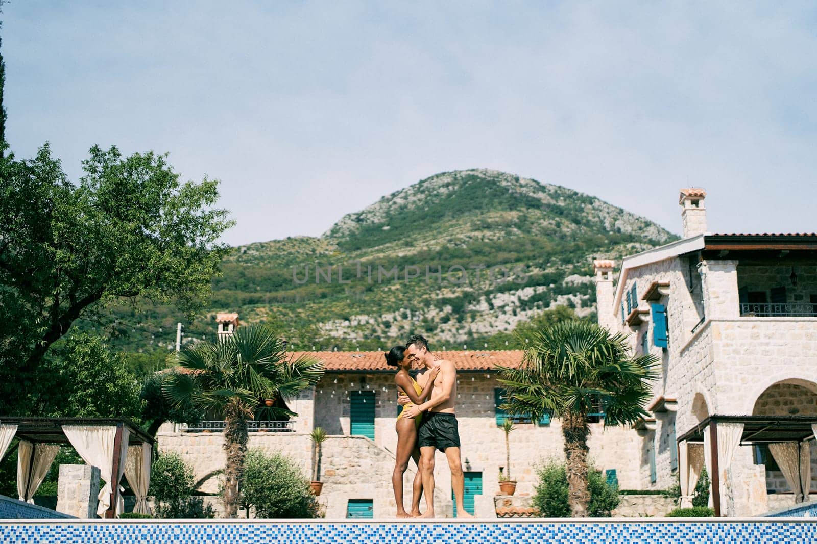 Man and woman hug while standing by the pool in front of an ancient villa by Nadtochiy
