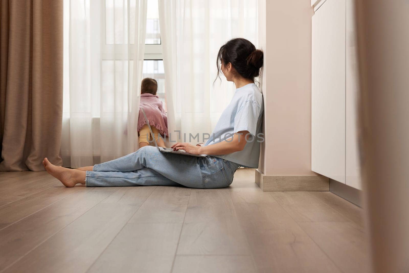 woman sitting on the floor and working on laptop with her little child girl at home by erstudio