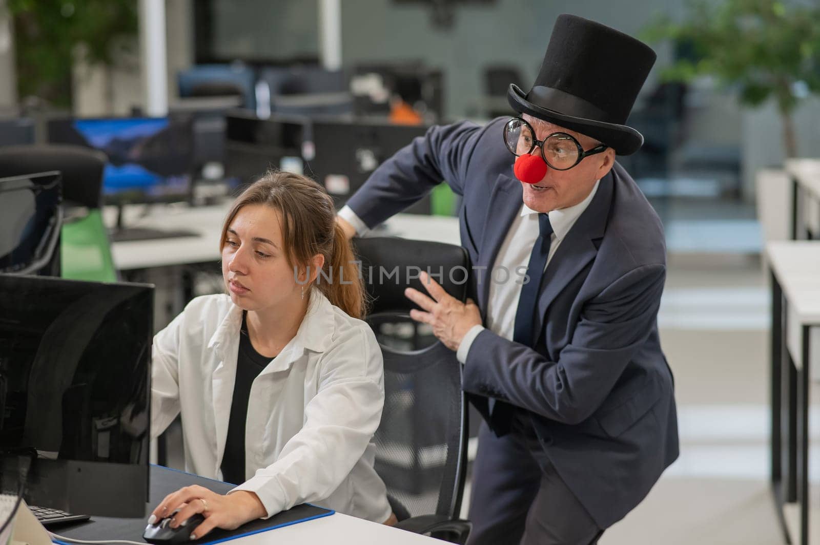 Caucasian woman works at the computer. Elderly man in clown costume in office. by mrwed54