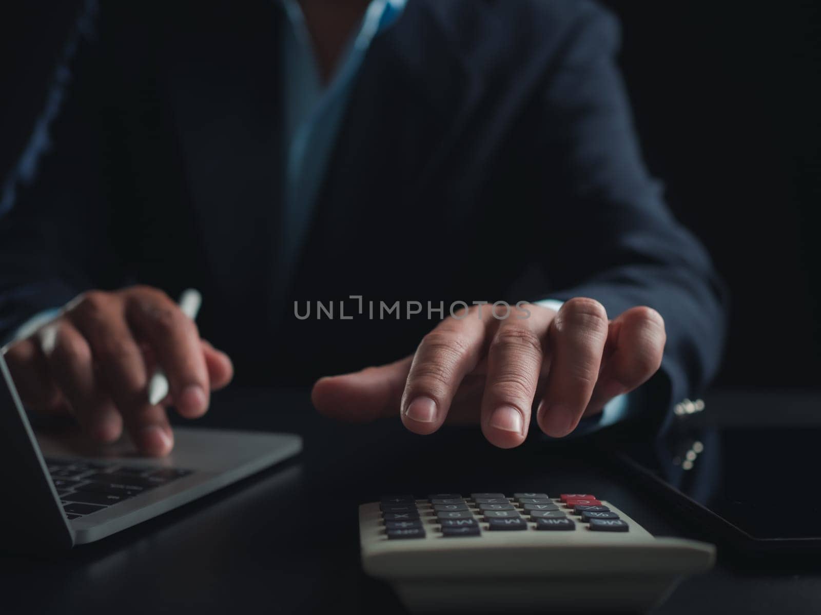 Businessman in black suit working on laptop computer and tablet and calculator, Hand Hands touch on tablet at office with dark background, Online working, Close up, Copy space.