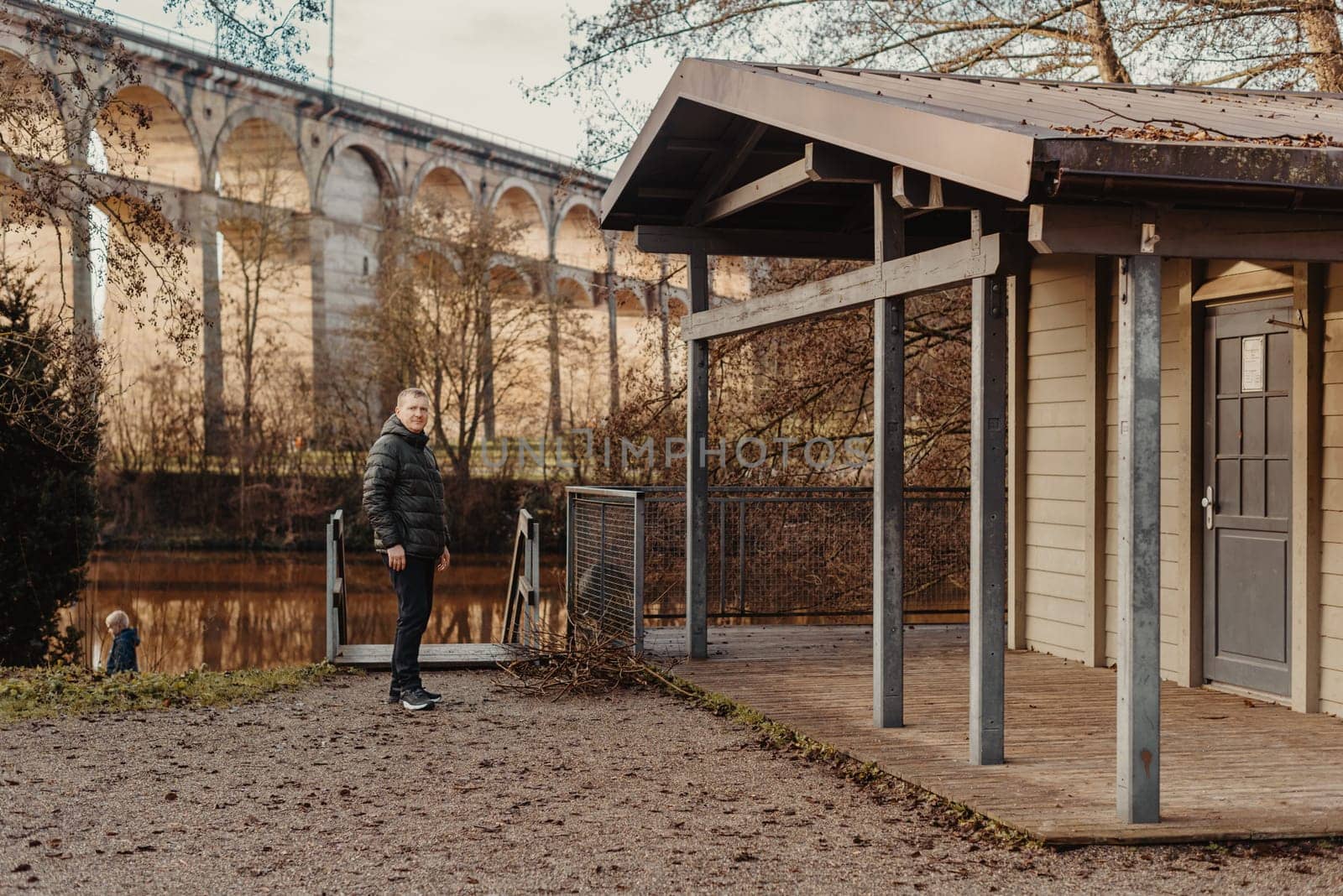 Timeless Elegance: 40-Year-Old Man in Stylish Jacket by Neckar River and Historic Bridge in Bietigheim-Bissingen, Germany. Experience the allure of seasons as a charismatic 40-year-old man stands gracefully by the enchanting Neckar River, adorned in a sophisticated jacket, against the backdrop of historic bridge pillars in the charming city of Bietigheim-Bissingen, Germany. This captivating image seamlessly blends timeless elegance with the scenic beauty of autumn or winter, creating a picturesque scene of urban exploration and mature style in the heart of German heritage