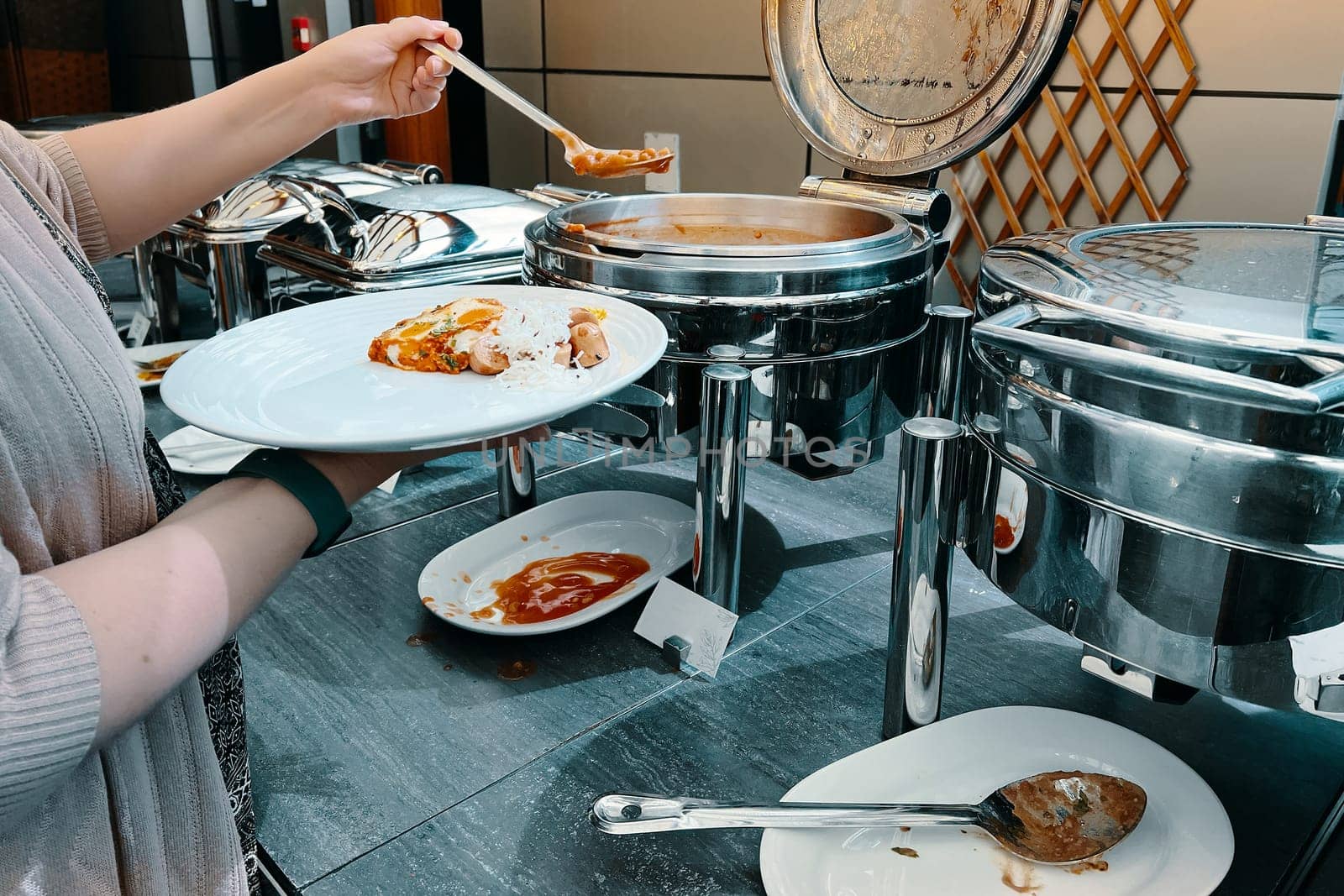 In the hotel, a lady is filling her plate at the buffet with food from a warming machine.