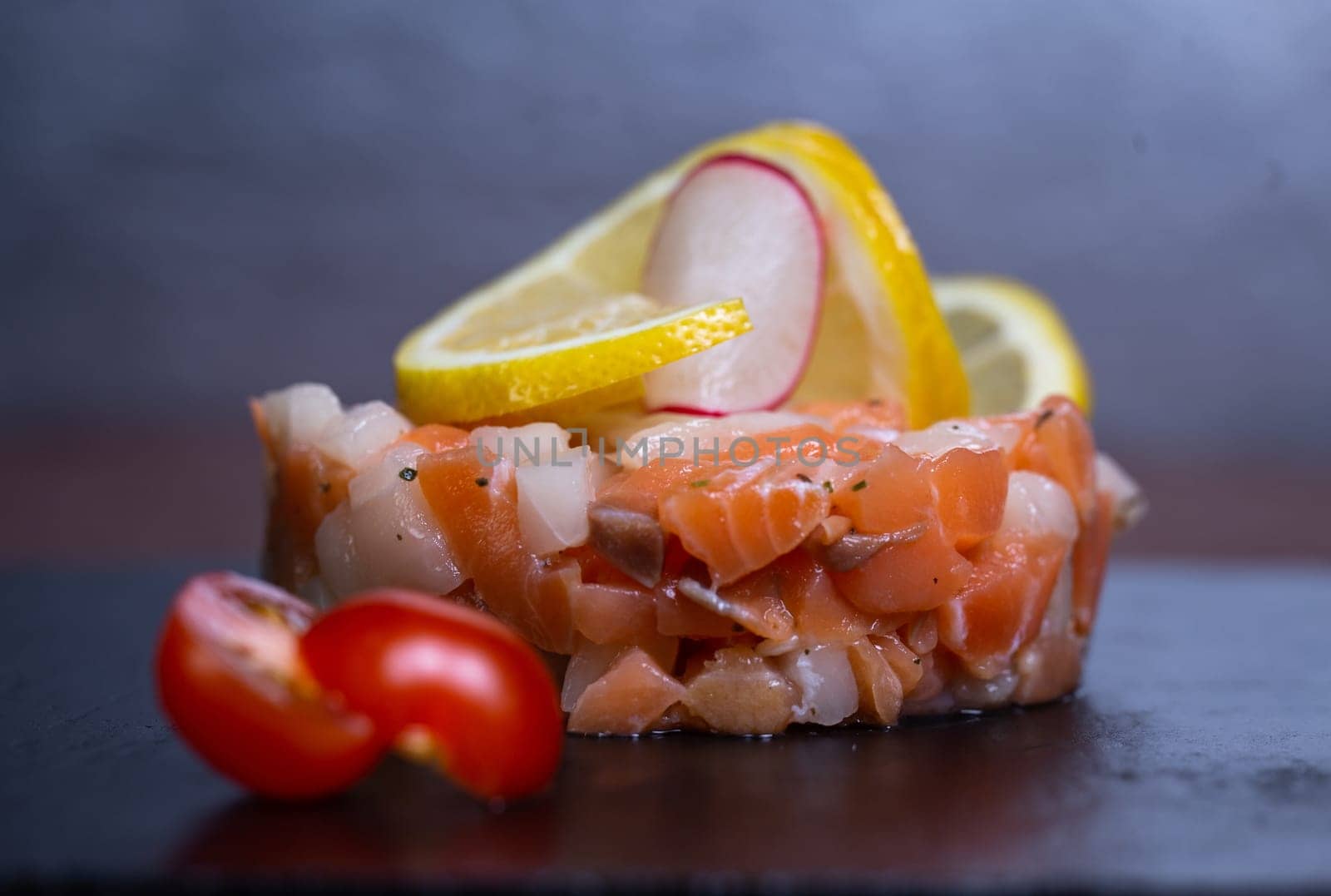 Savor a delightful salmon tartare with lemon, radish, and cherry tomato on a stylish slate plate for a burst of flavors and textures.