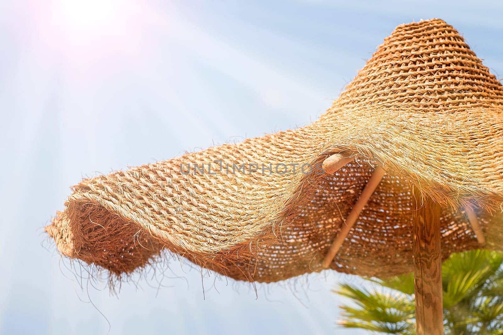 Wicker sun umbrella looks like a hat against blue sky and sun rays.