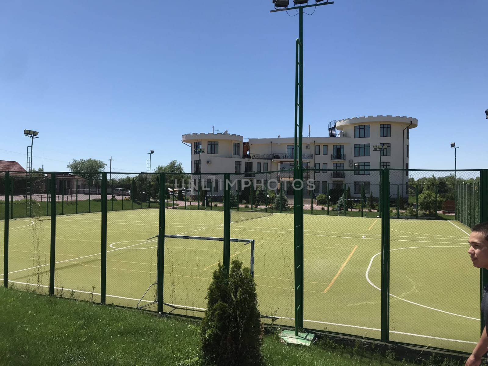 An outdoor soccer field with fake grass and a hotel with a unique architecture in the background under a clear blue sky.