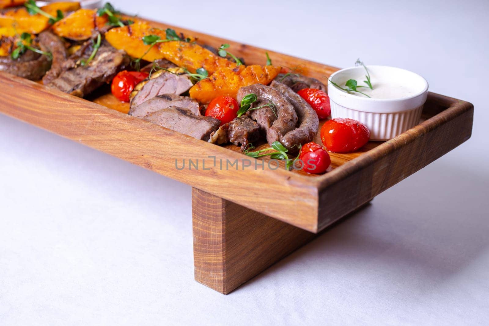 A long wooden platter filled with an assortment of meats and vegetables, including steak, sausage, chicken, potatoes, carrots, and peppers by Pukhovskiy