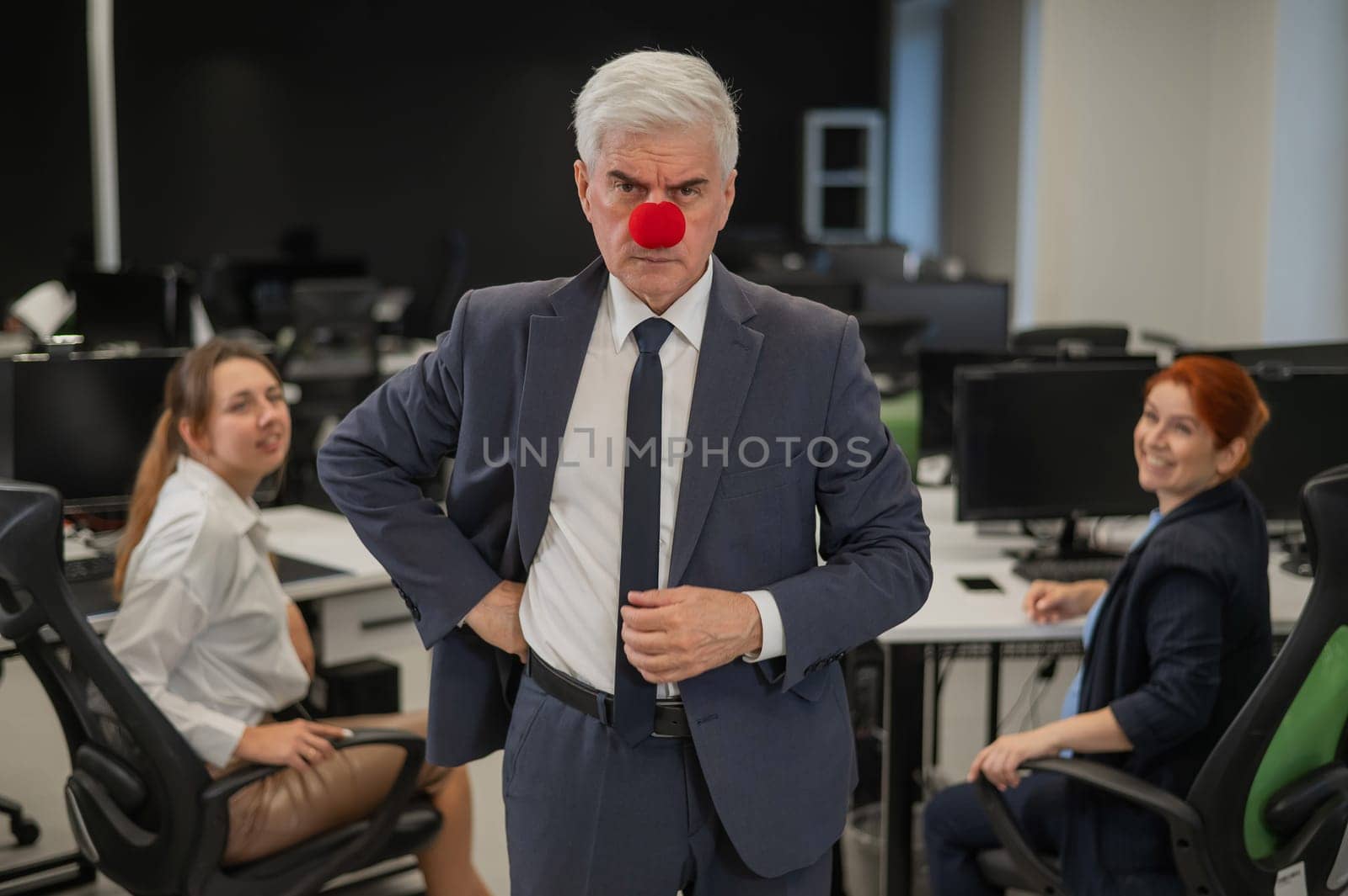 Two Caucasian women look at a serious elderly man with a clown nose in the office
