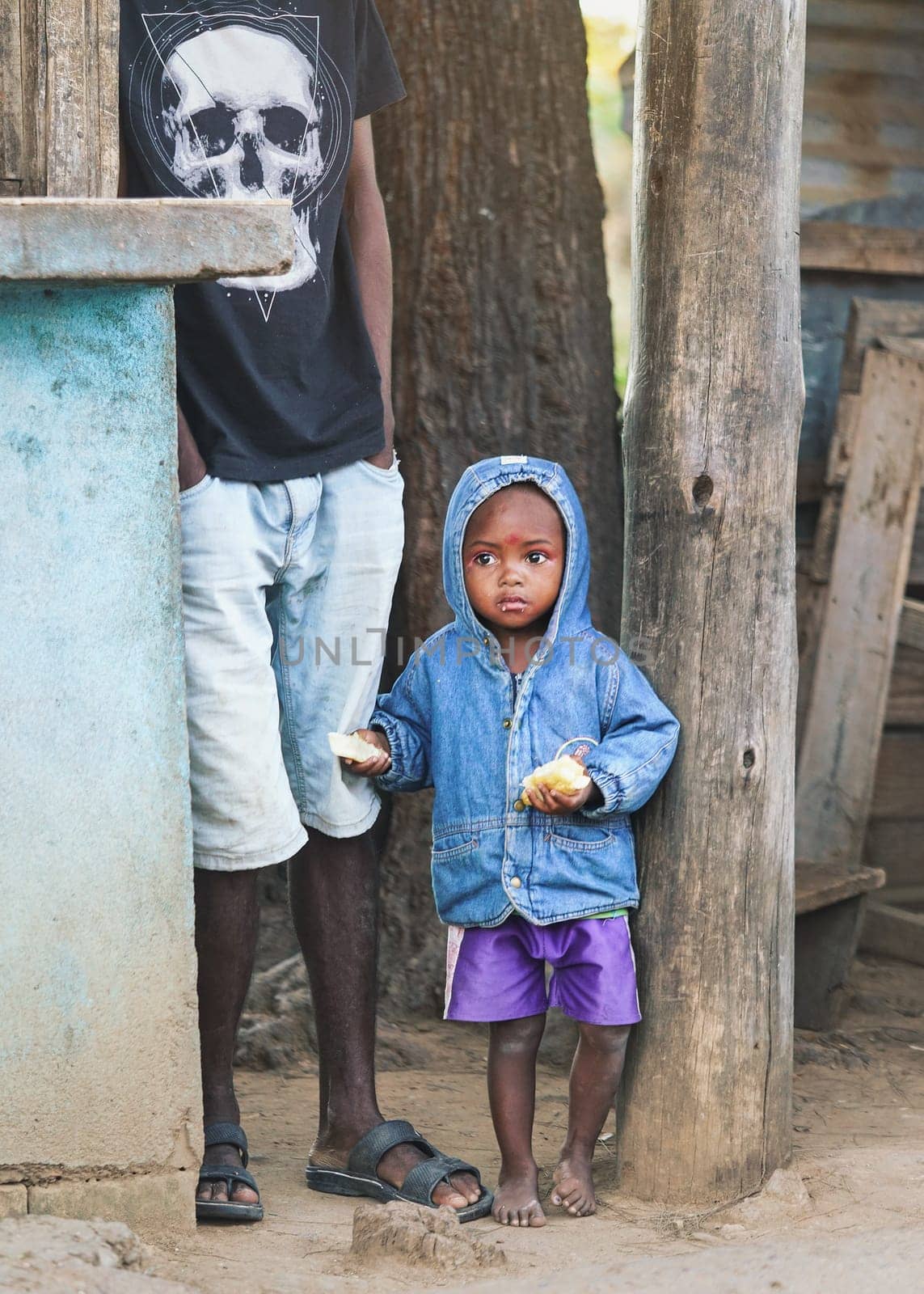 Ranohira, Madagascar - April 29, 2019: Unknown small Malagasy boy kid in blue denim jacked and shorts standing next to his father, holding bread in both hands. 