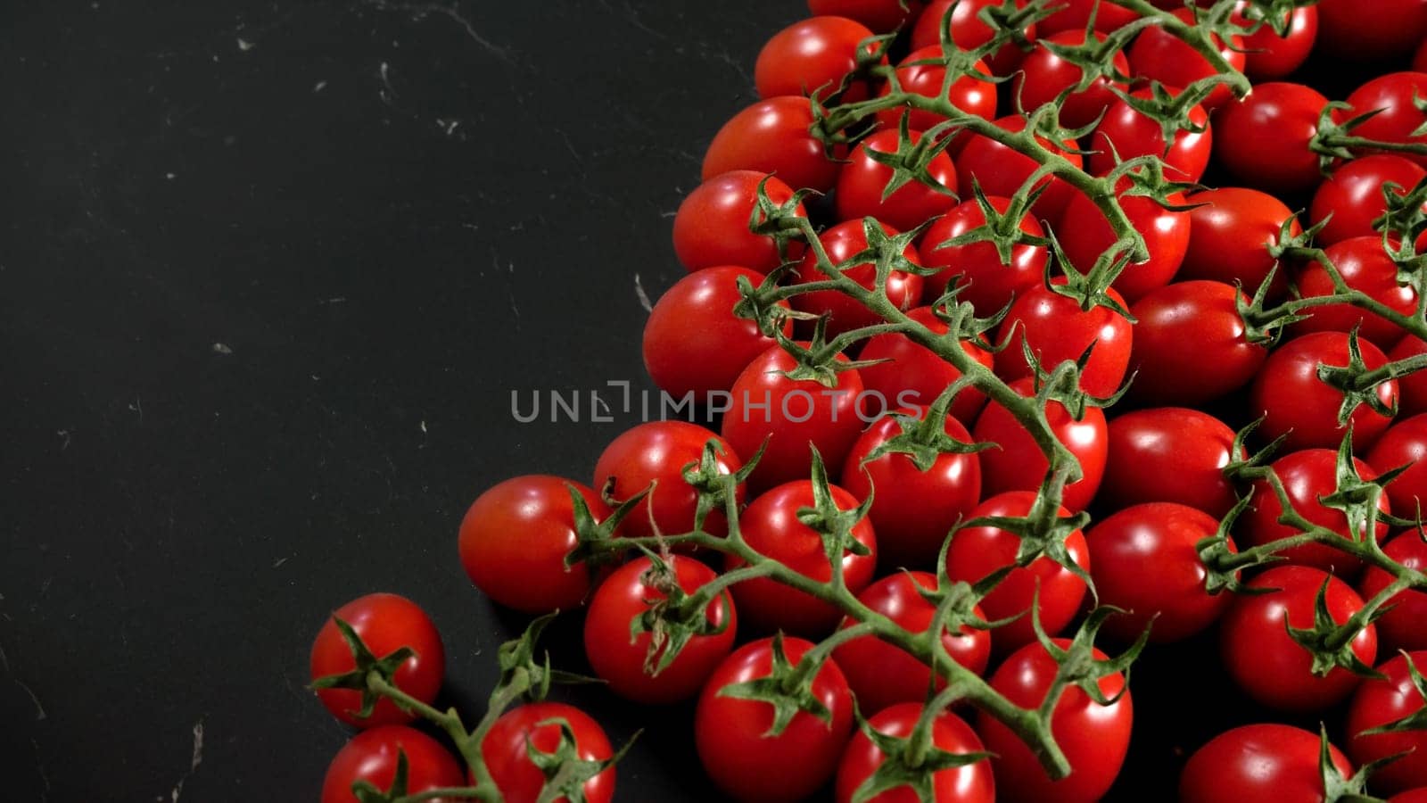 Bright red cherry tomatoes with green leaves on black marble like board, space for text left side