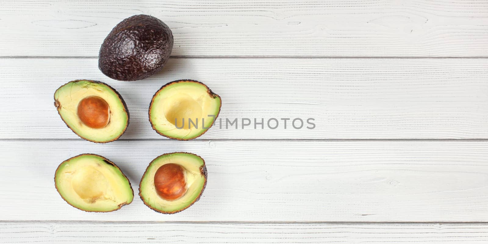 Ripe avocado halves and one whole fruit arranged on white boards desk, view from above - wide photo with space for text right side