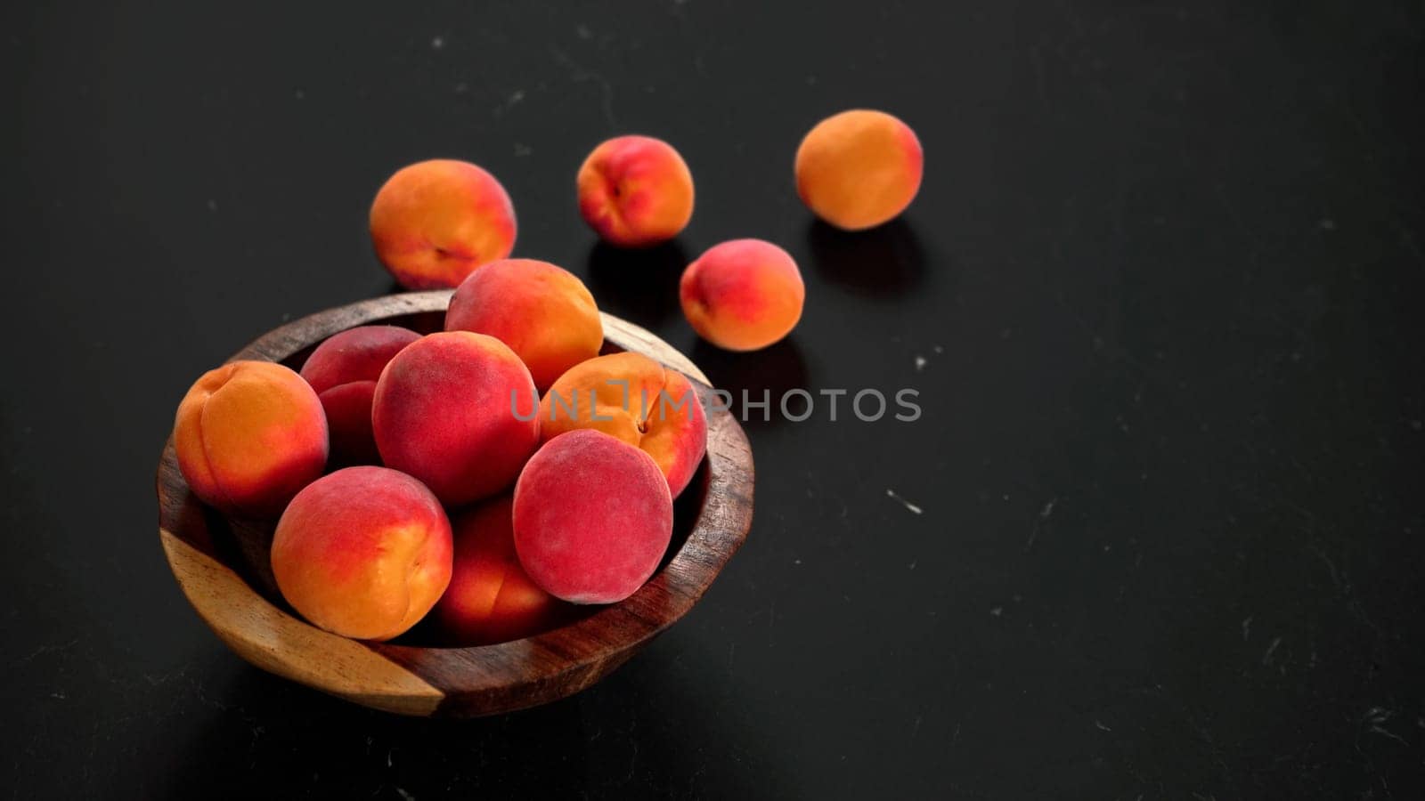 Apricots in small wooden bowl, some spilled on black table, space for text right side