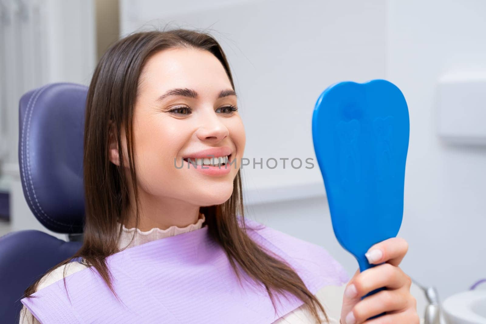 Happy brunette woman using mirror while looking at her teeth after dental treatment.