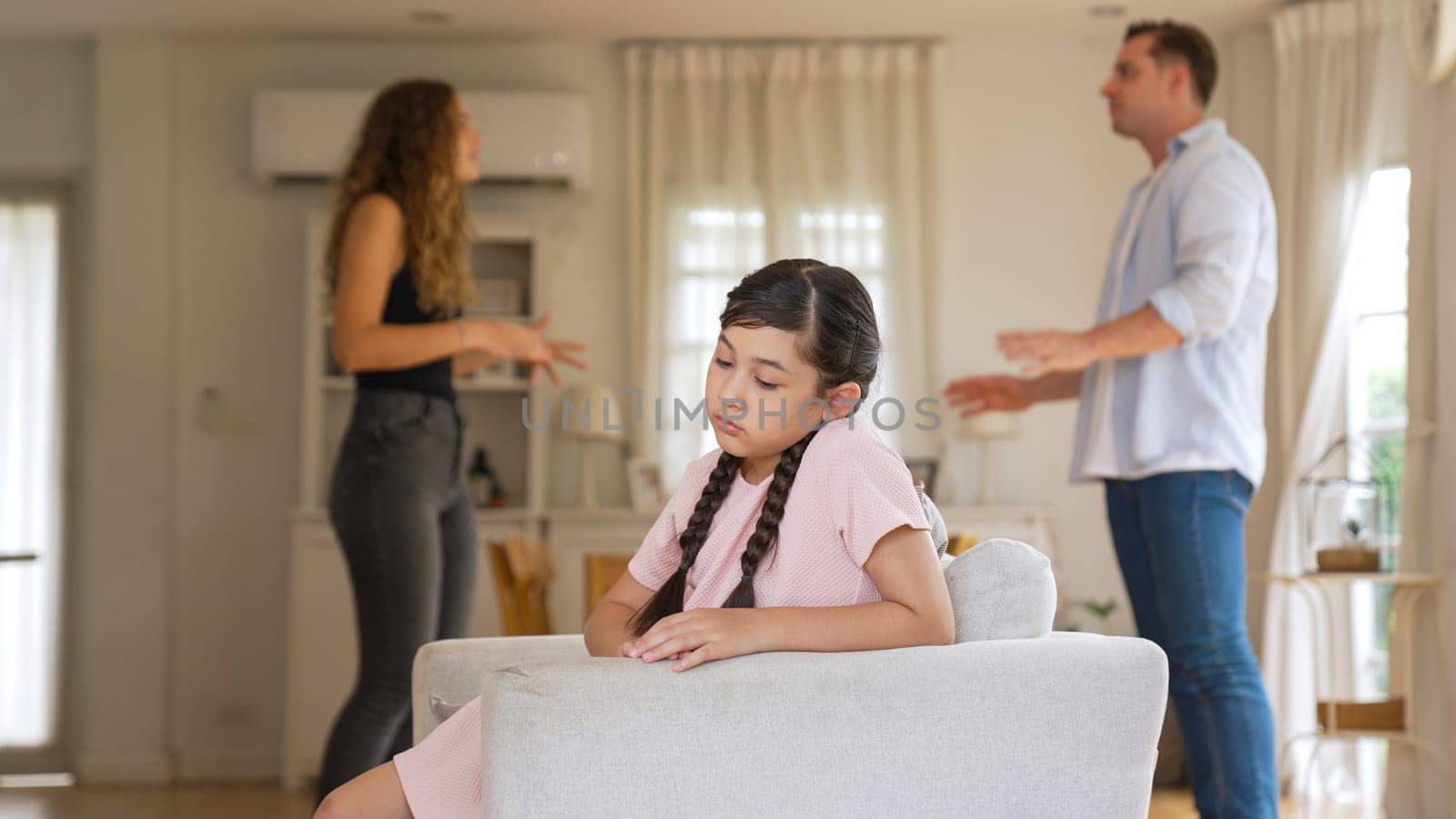 Annoyed and unhappy young girl sitting on sofa trapped in middle of tension by her parent argument in living room. Unhealthy domestic lifestyle and traumatic childhood develop to depression Synchronos