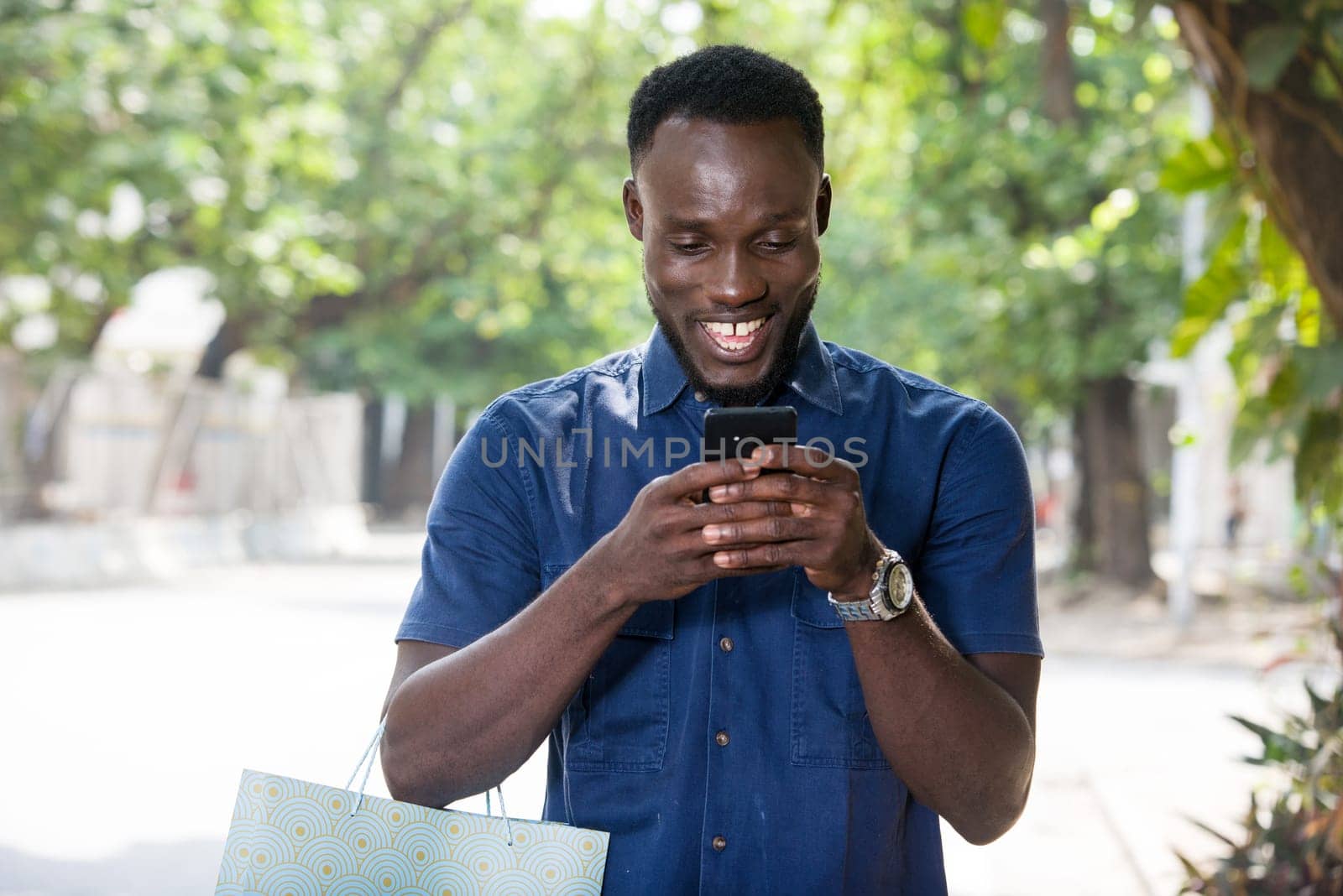 young man standing outdoors with shopping bag going to look at mobile phone while smiling.