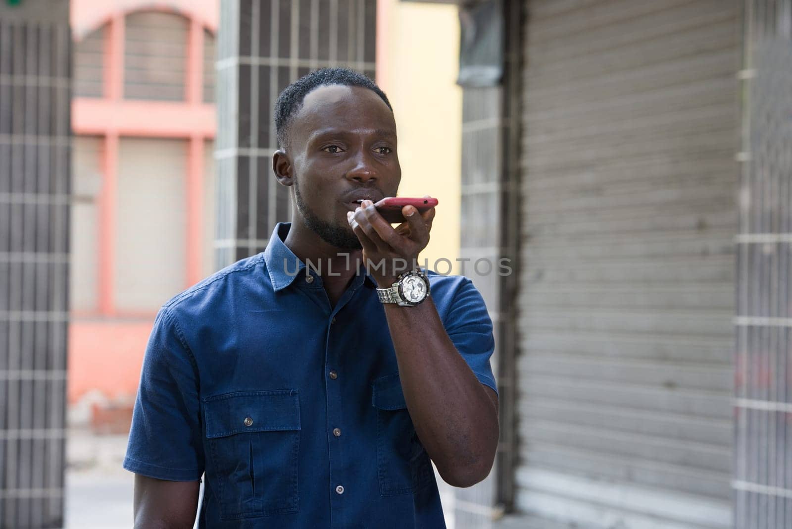 Outdoor portrait of attractive young african man, holding an empty smart-phone, speaks using voice command, feeling happy and surprised. Human emotions, concept of facial expression