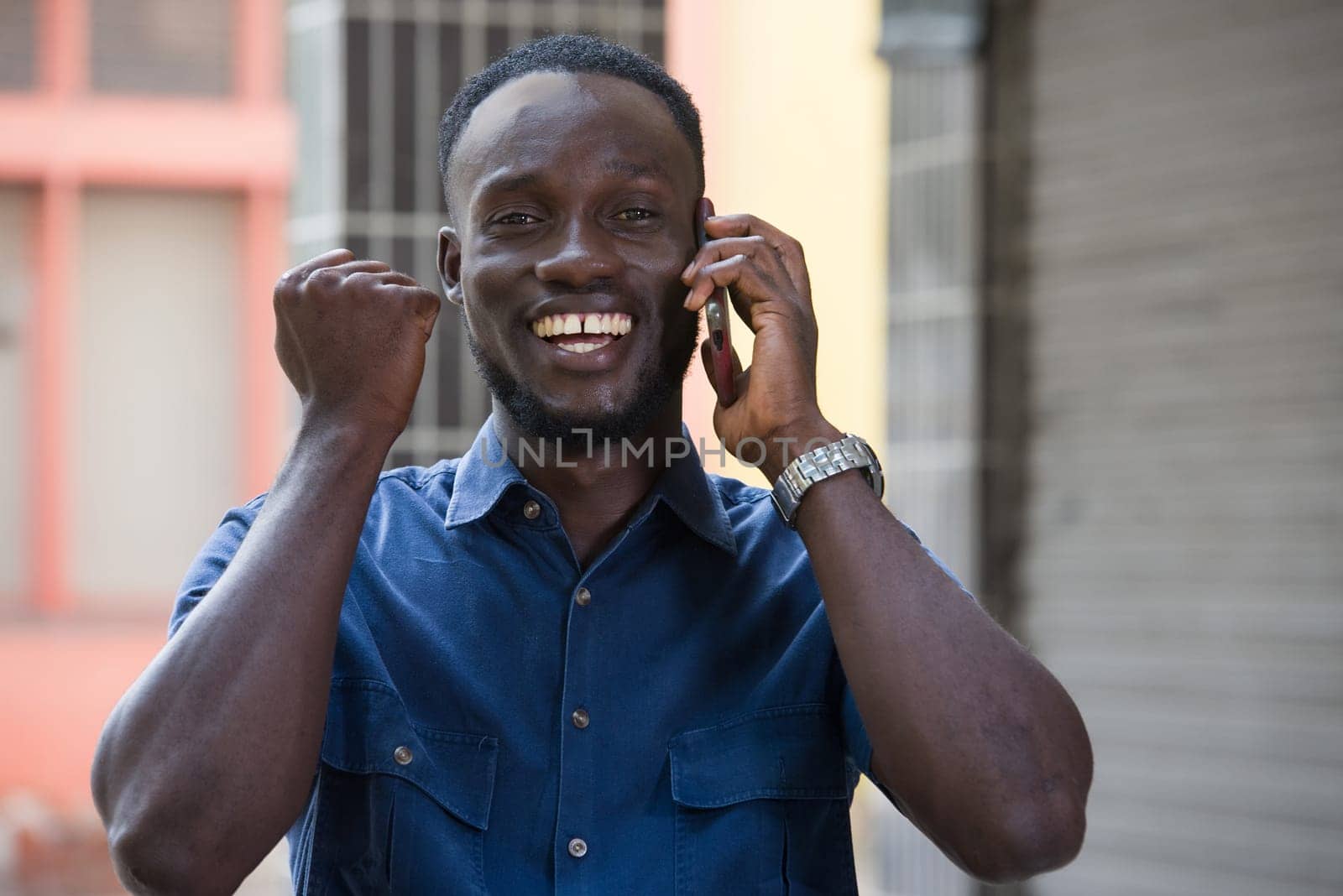 Photo of handsome excited man expressing surprise on face and gesturing while talking on the phone standing outside