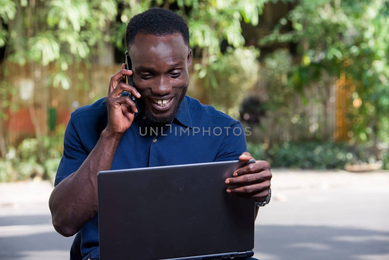 young man sitting outdoors going to communicate with mobile phone and looking at laptop smiling.