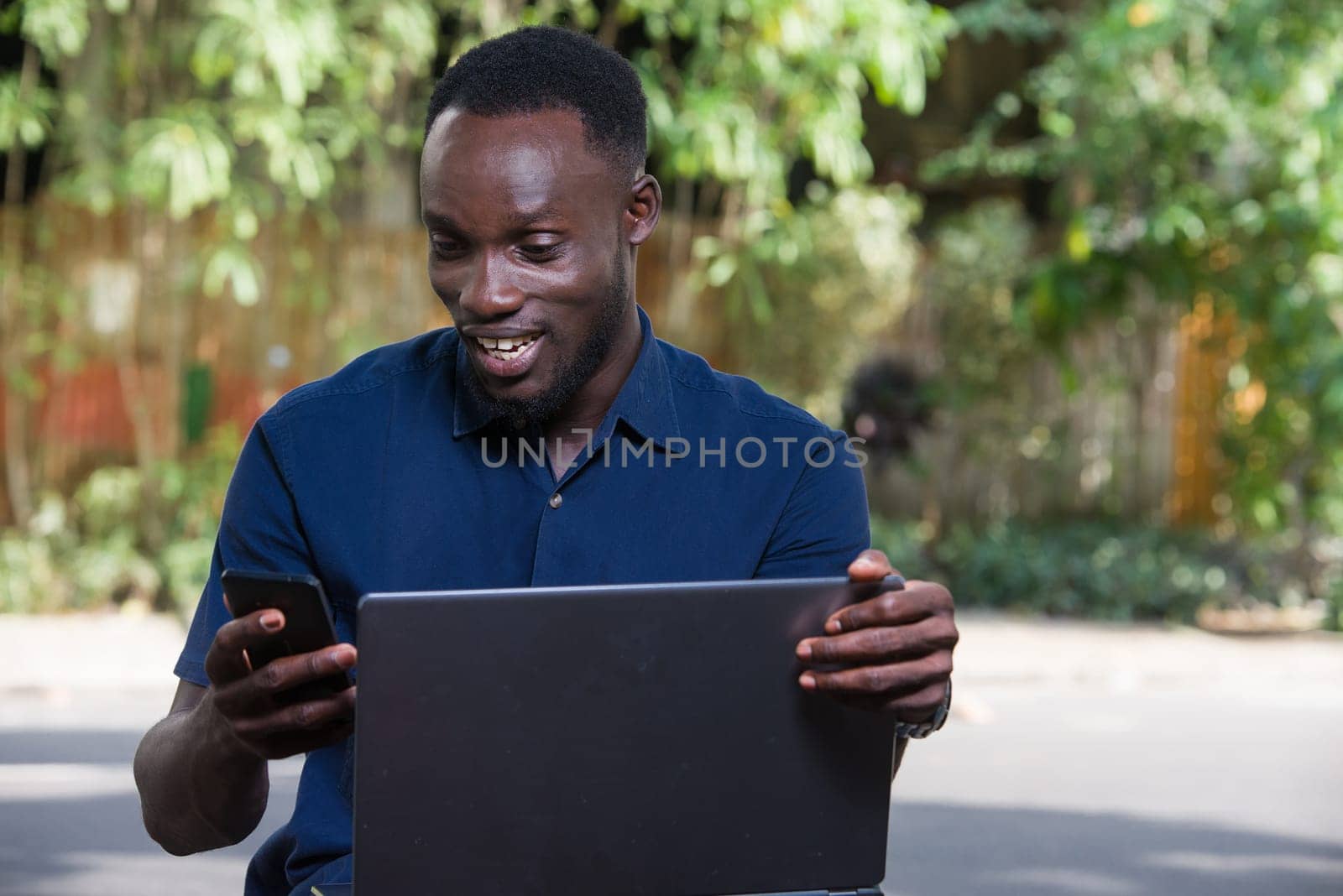 young man sitting outdoors with laptop watching mobile phone while smiling.