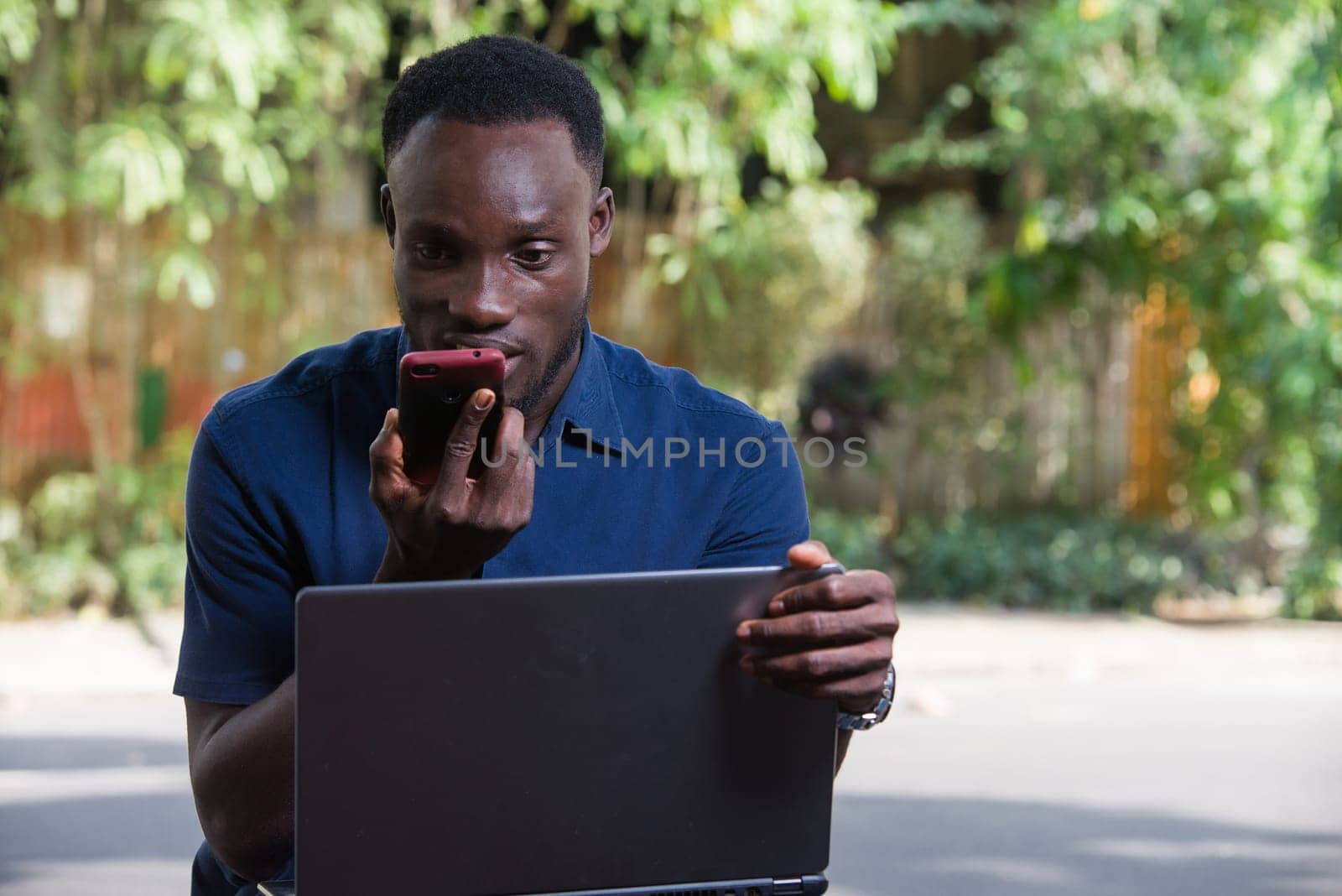 young man sitting outdoors with laptop watching mobile phone while smiling.