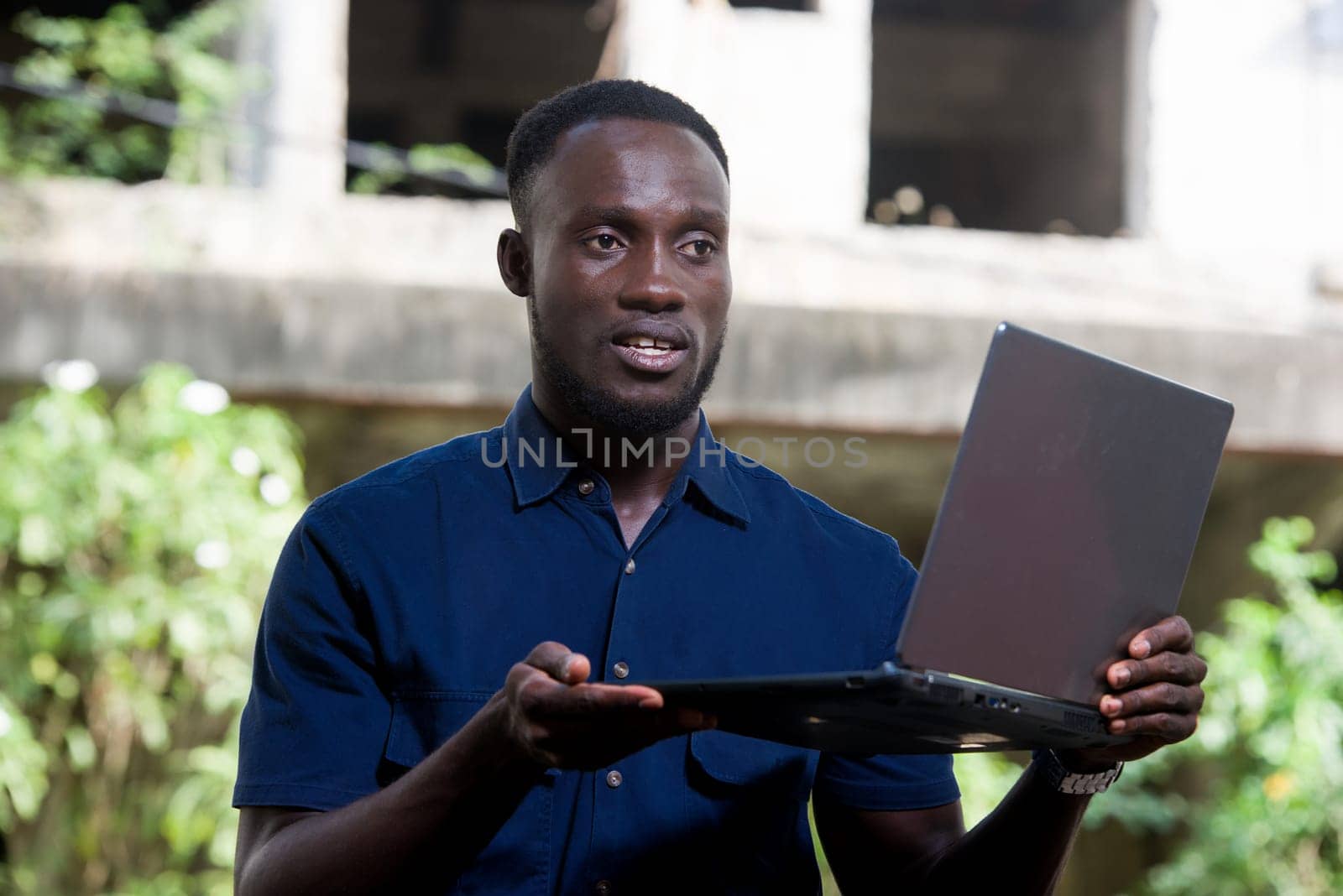 young man sitting outdoors presenting laptop while smiling.
