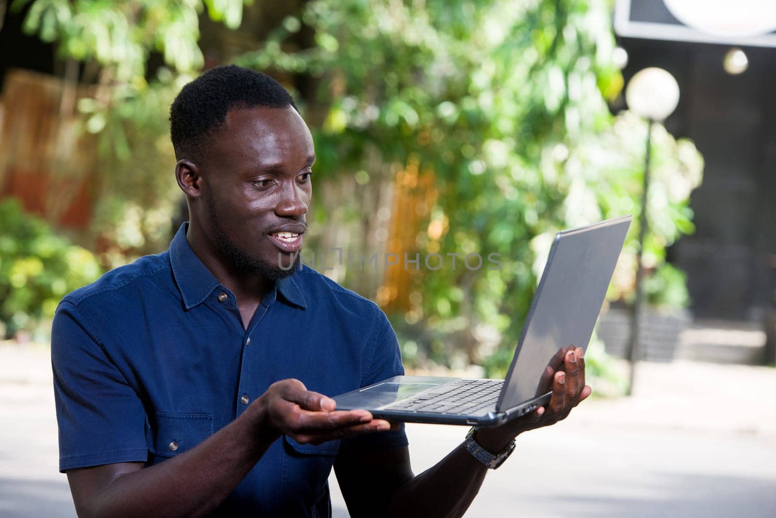 young man sitting outdoors presenting laptop while smiling.