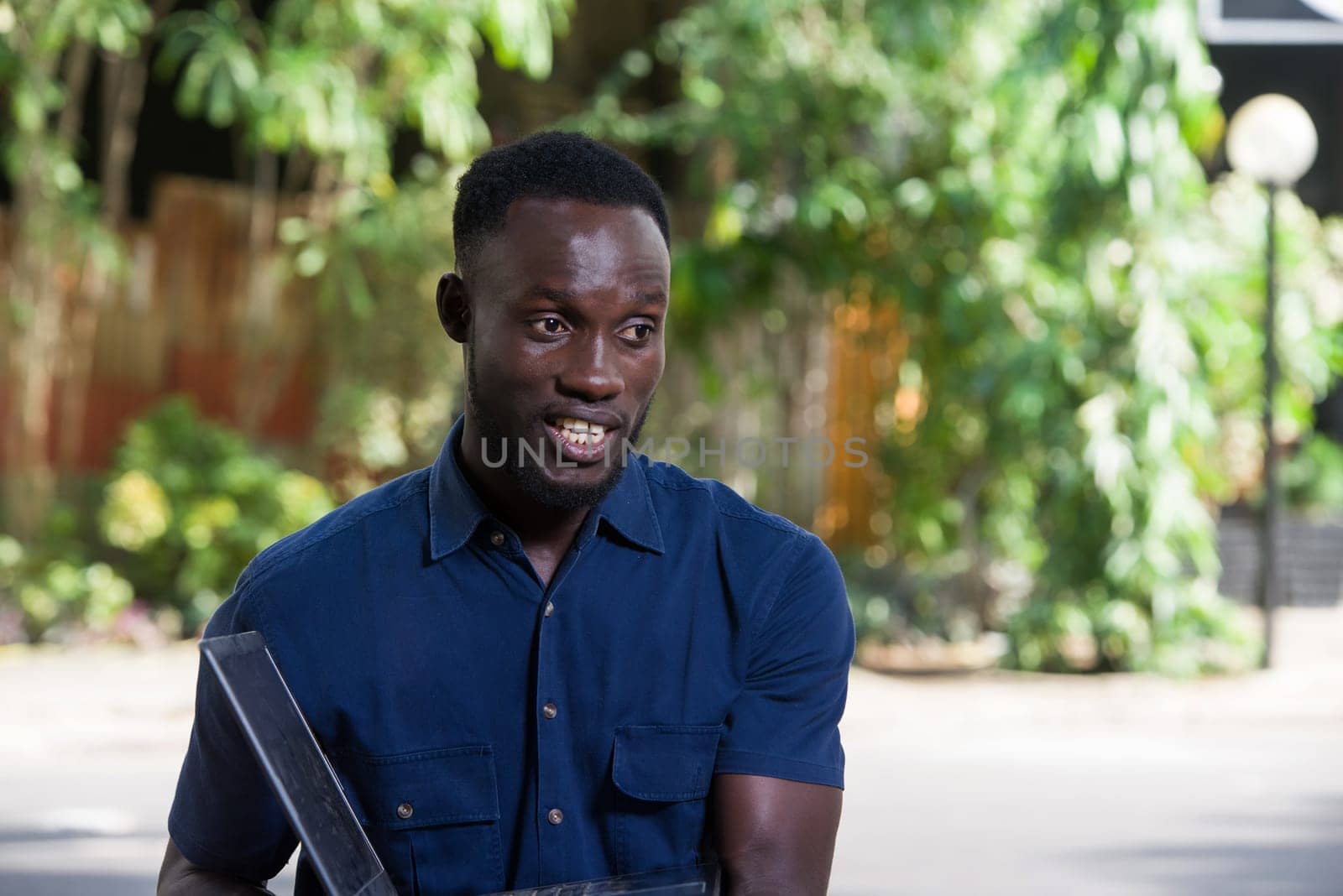 young man sitting outdoors presenting laptop while smiling.