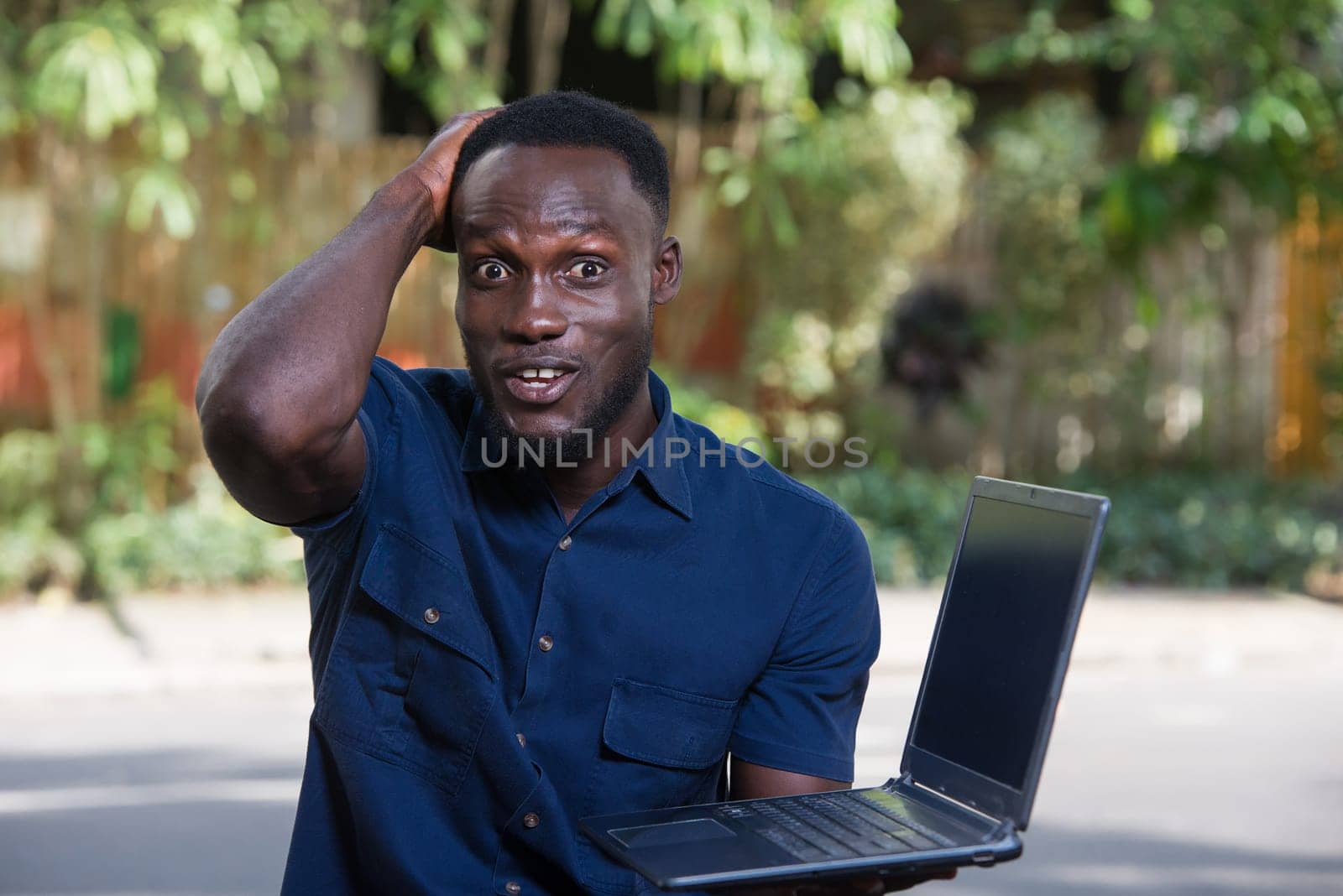 young man with laptop sitting outdoors looking at camera smiling.