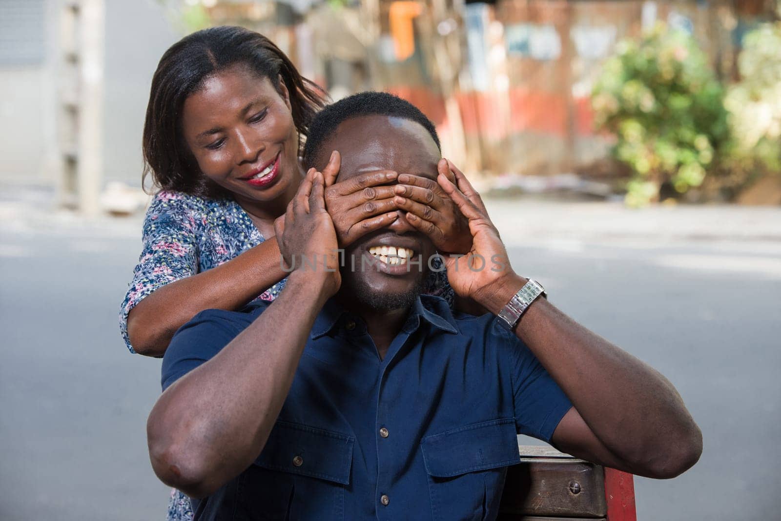 young woman standing outdoors playing with her boyfriend covering her eyes with her hand while smiling.