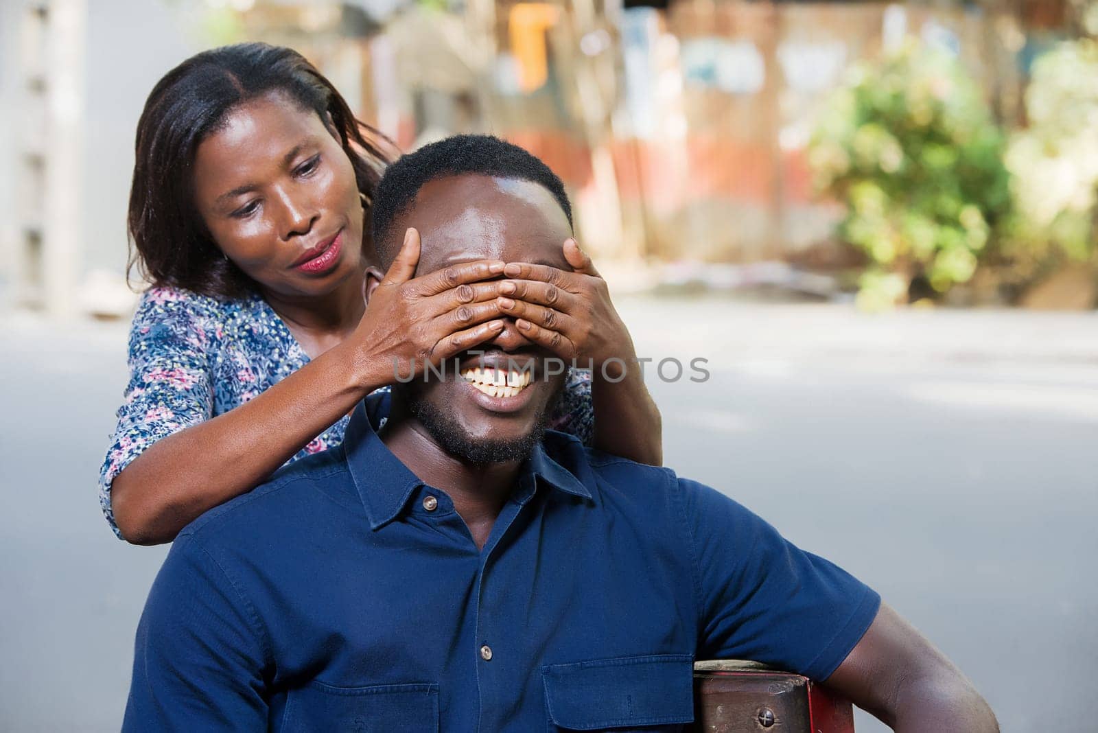 young woman standing outdoors playing with her boyfriend covering her eyes with her hand while smiling.