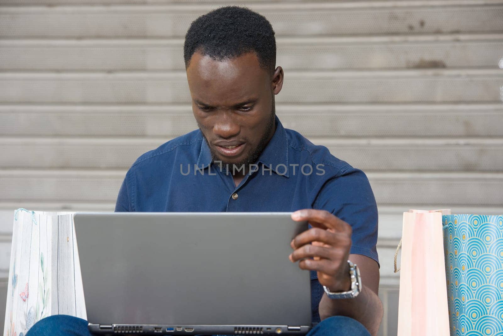 young man sitting on the floor with shopping bags watching laptop looking displeased.